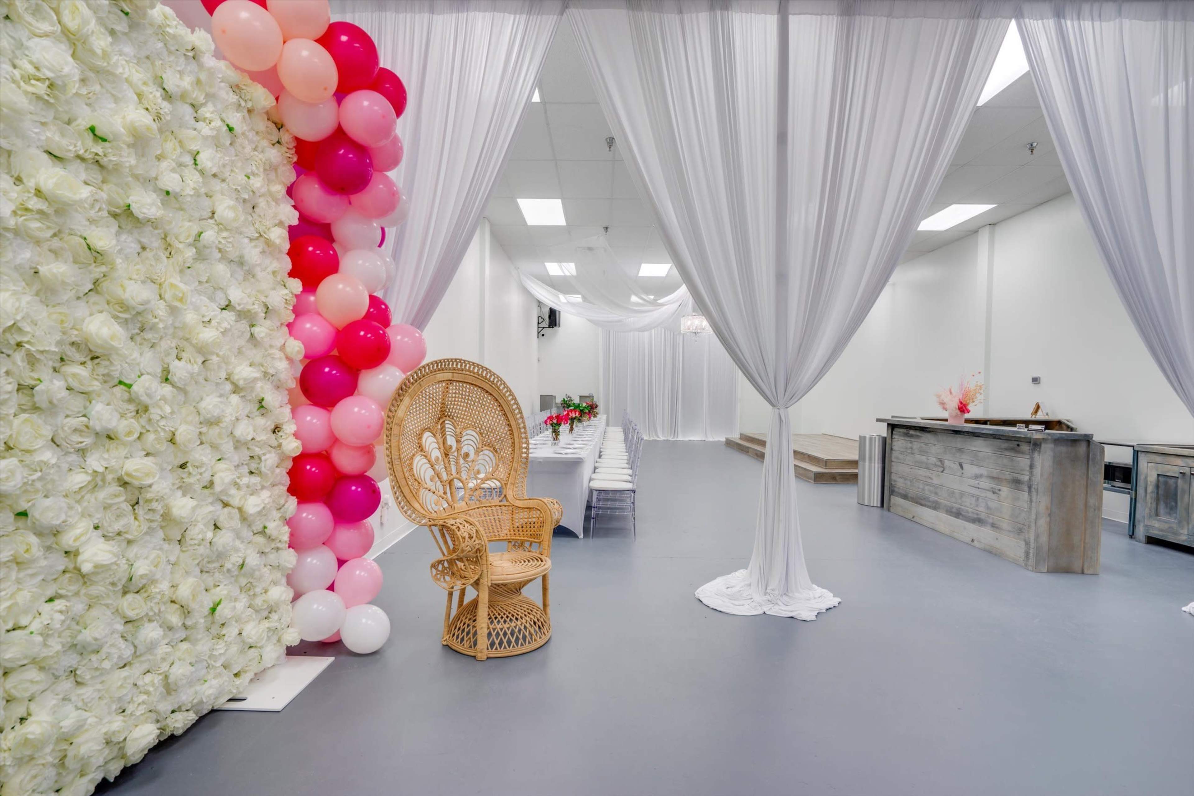 The image features a spacious event space with a flower wall, pink and white balloon decorations, a rattan chair, and tables set for a gathering.