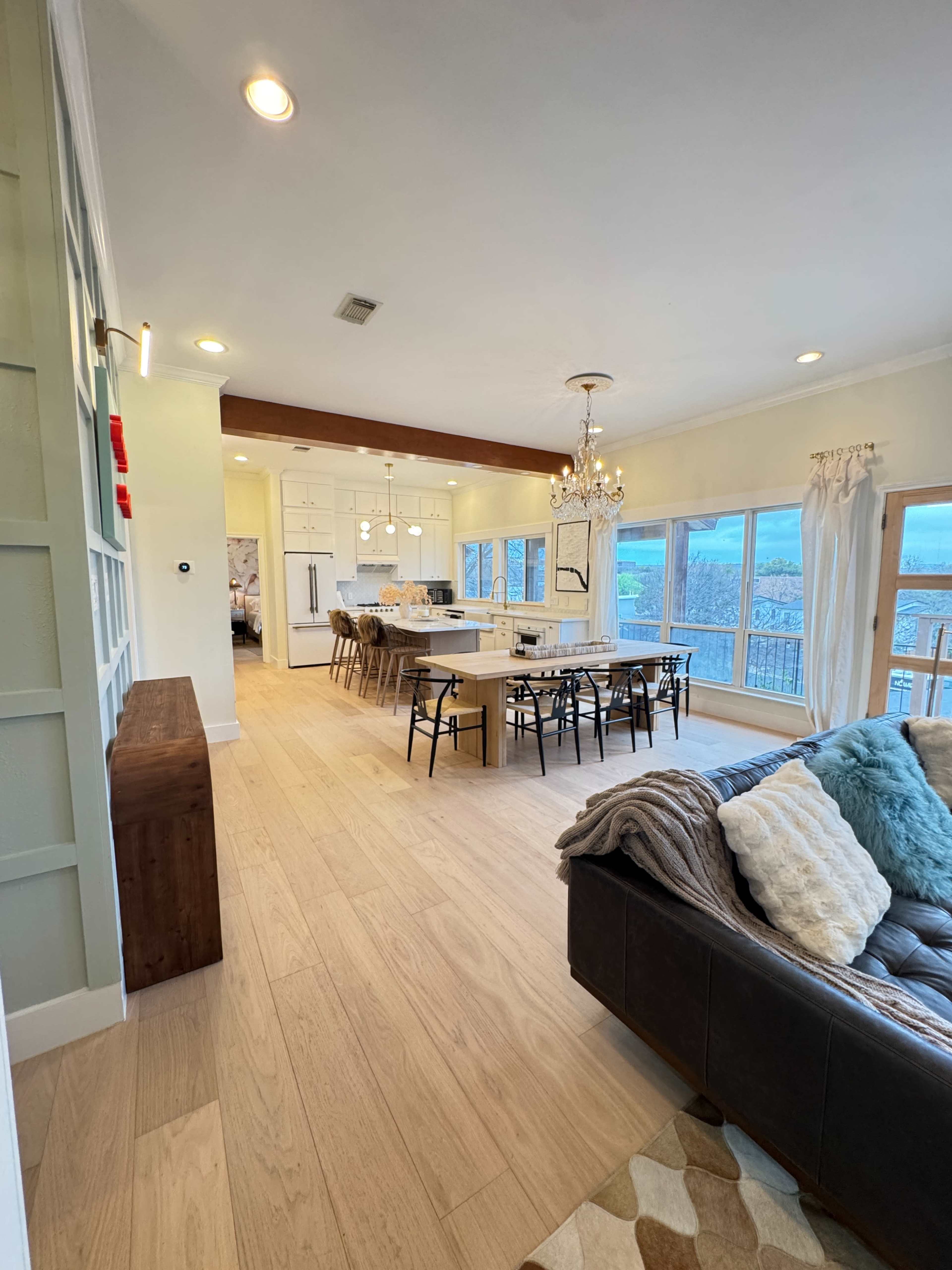 The image shows a spacious, modern living area that opens into a dining space with large windows offering a view of the outdoors.