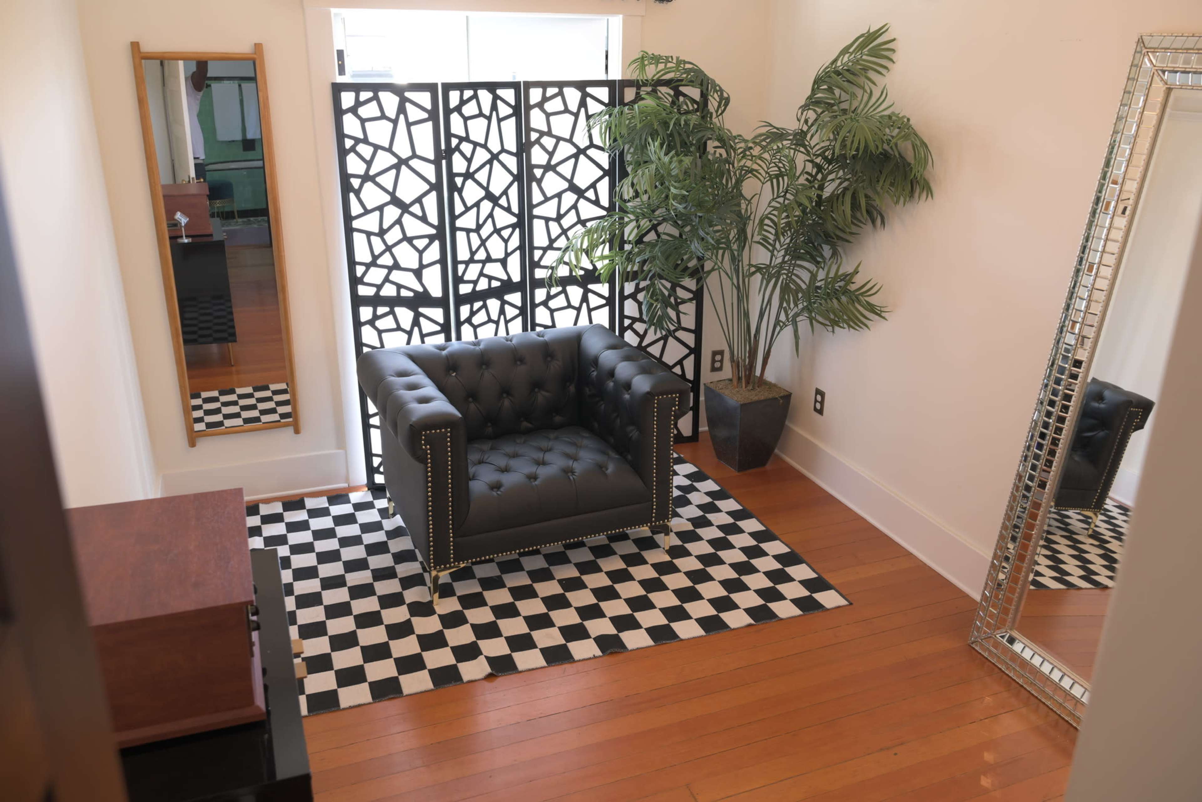 A room with a black leather tufted chair on a black-and-white checkered rug, a large mirror, and a decorative folding screen next to a potted plant.