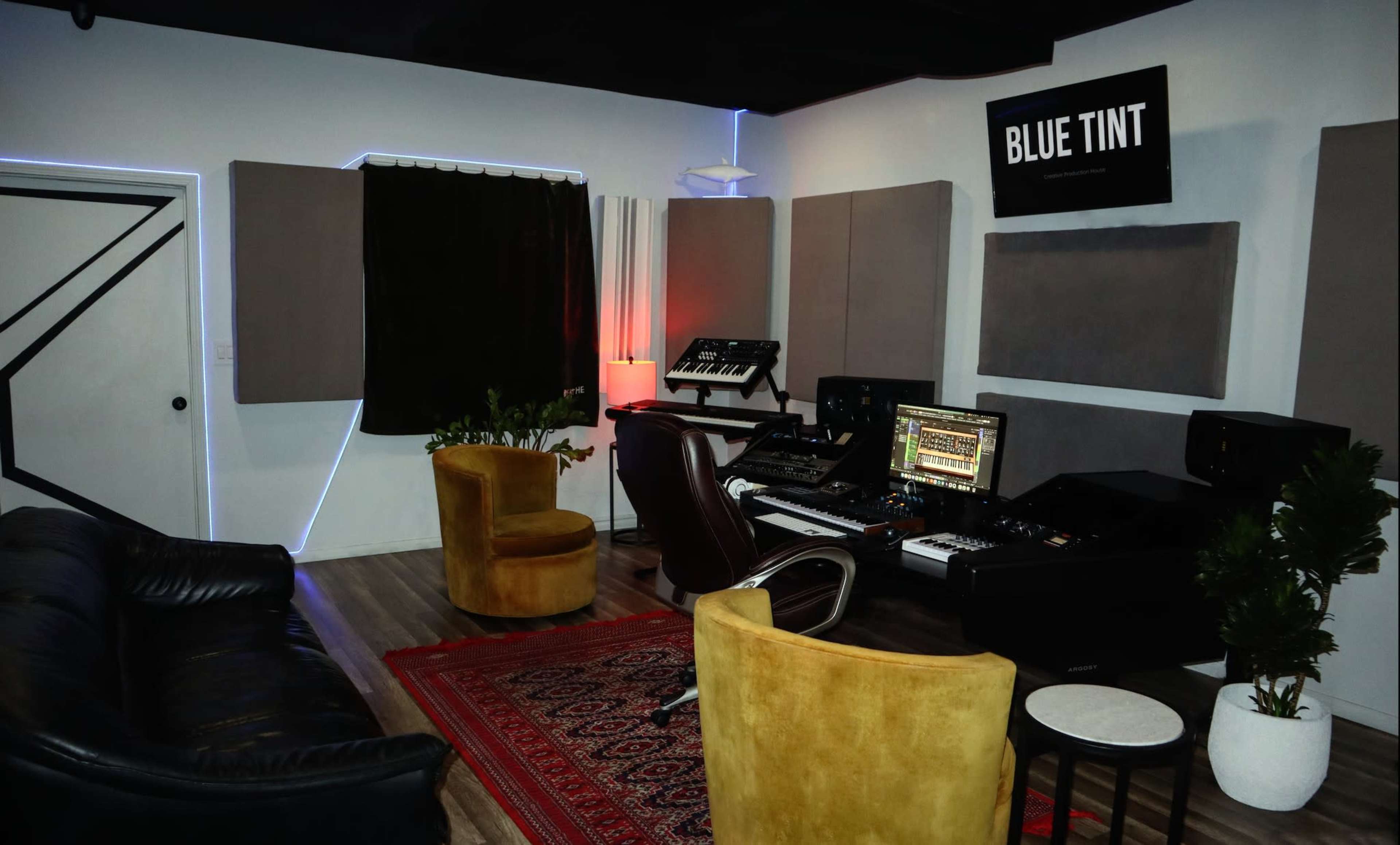 The image shows a music production studio equipped with synthesizers, a mixing console, and comfortable seating under soft lighting.
