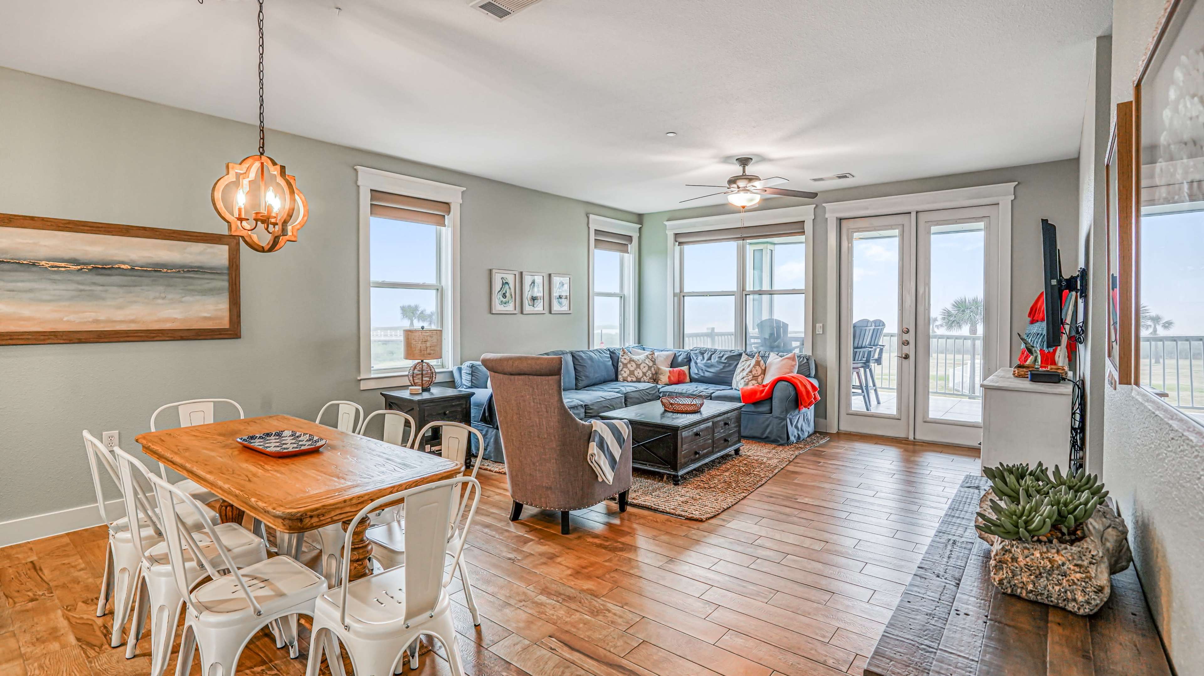 A bright, spacious living area features a dining table with metal chairs, a sectional sofa, and large windows overlooking a beach view.