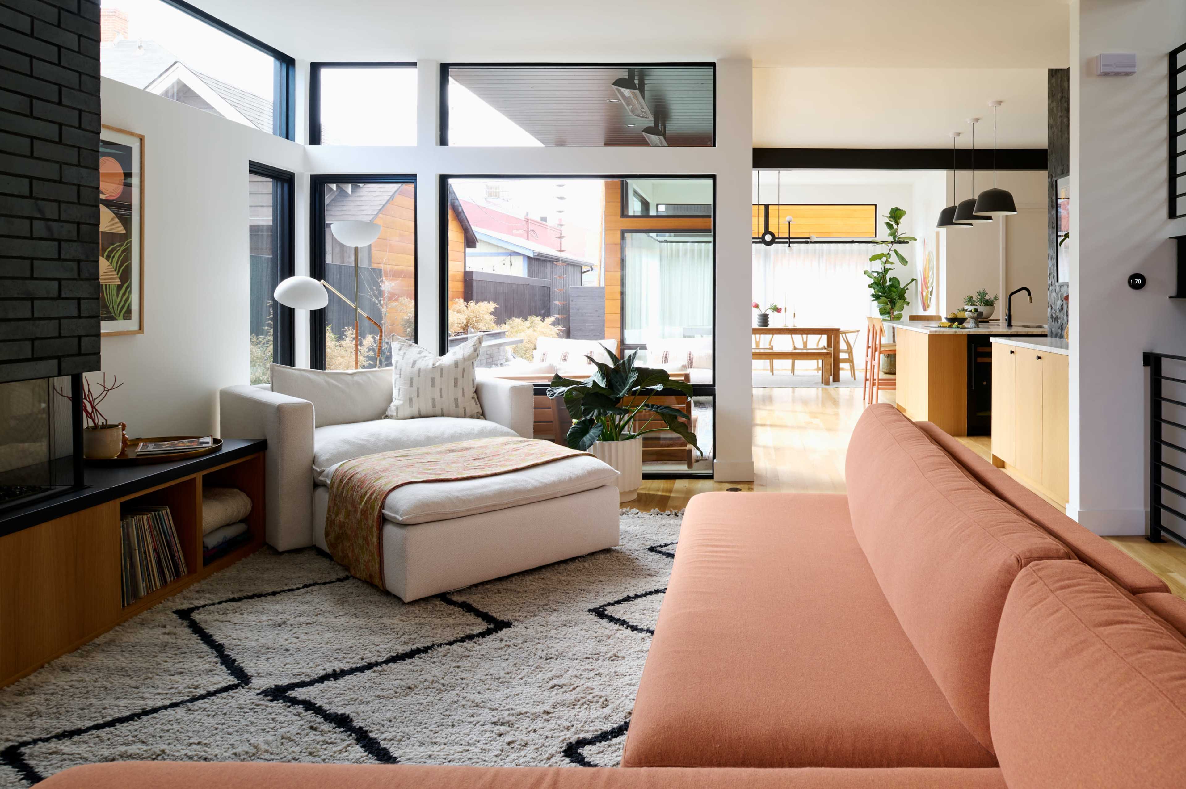 The image shows a modern living room with large windows, a sectional sofa, a lounge chair, and an open layout leading to a dining area and kitchen.