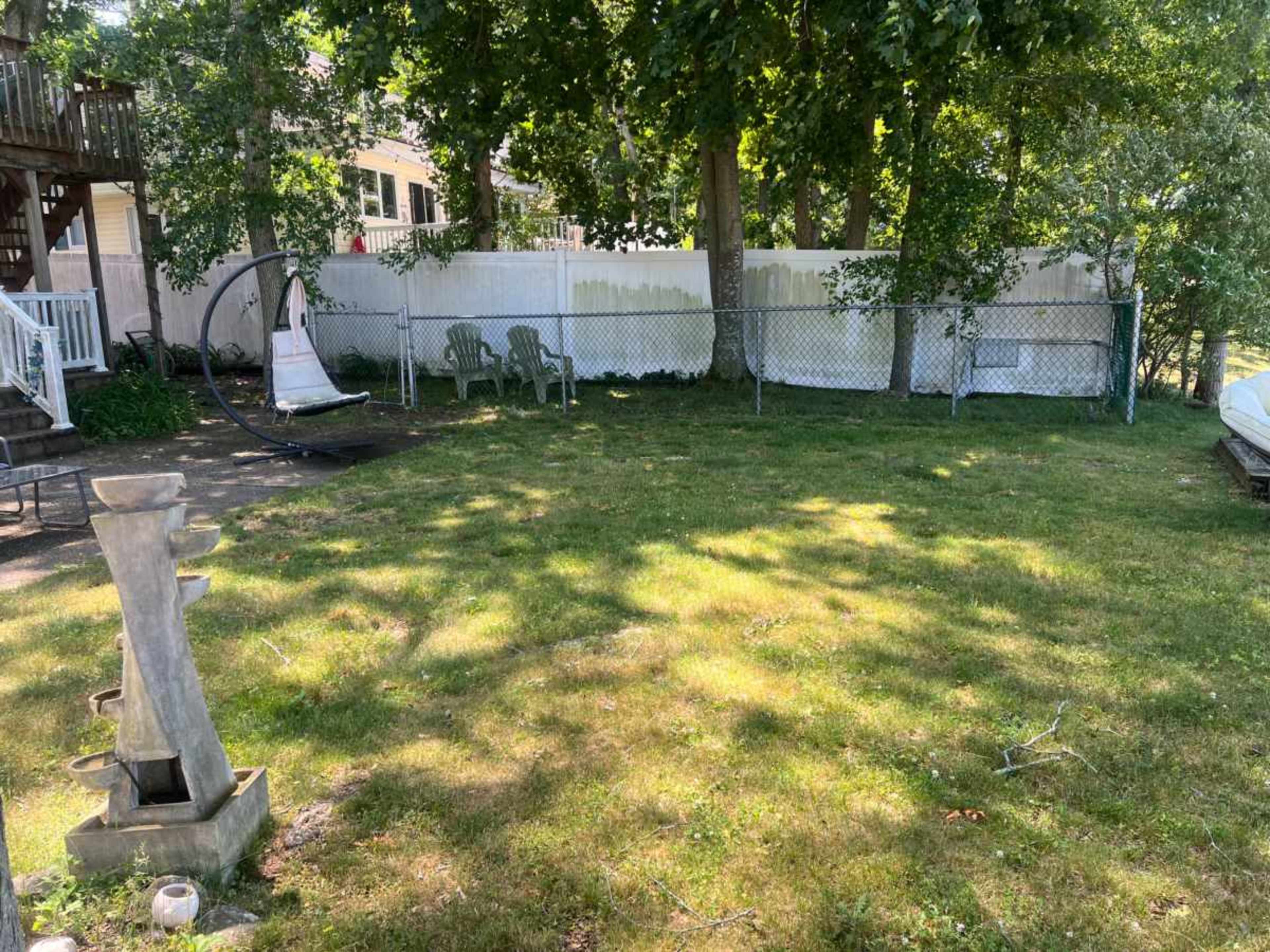 The image shows a grassy backyard with a fenced area, a few plastic chairs, and a stone fountain in the foreground.