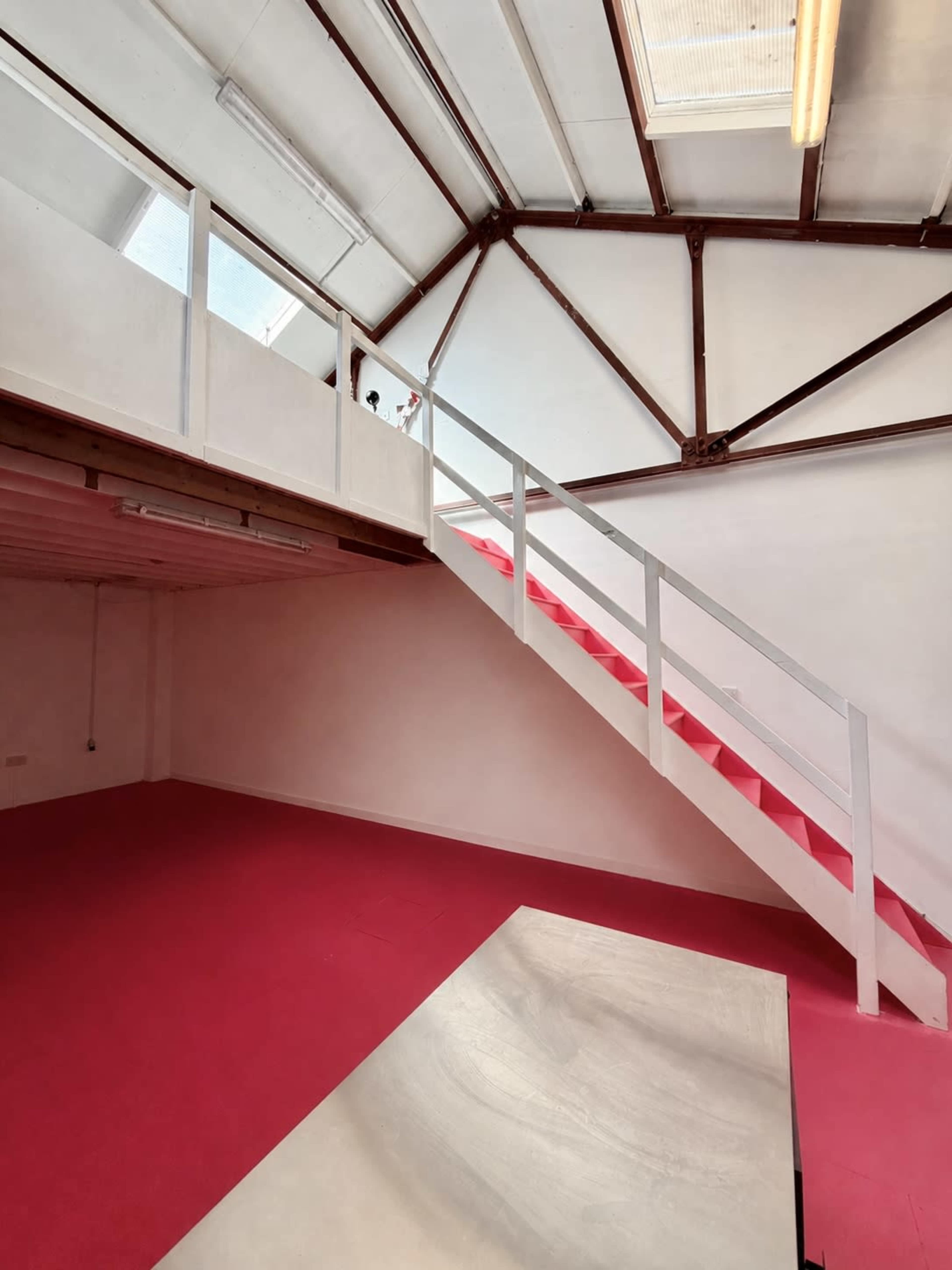 The image shows an indoor space with a red carpeted floor and a staircase leading to a loft area, featuring a wooden railing and large windows above.