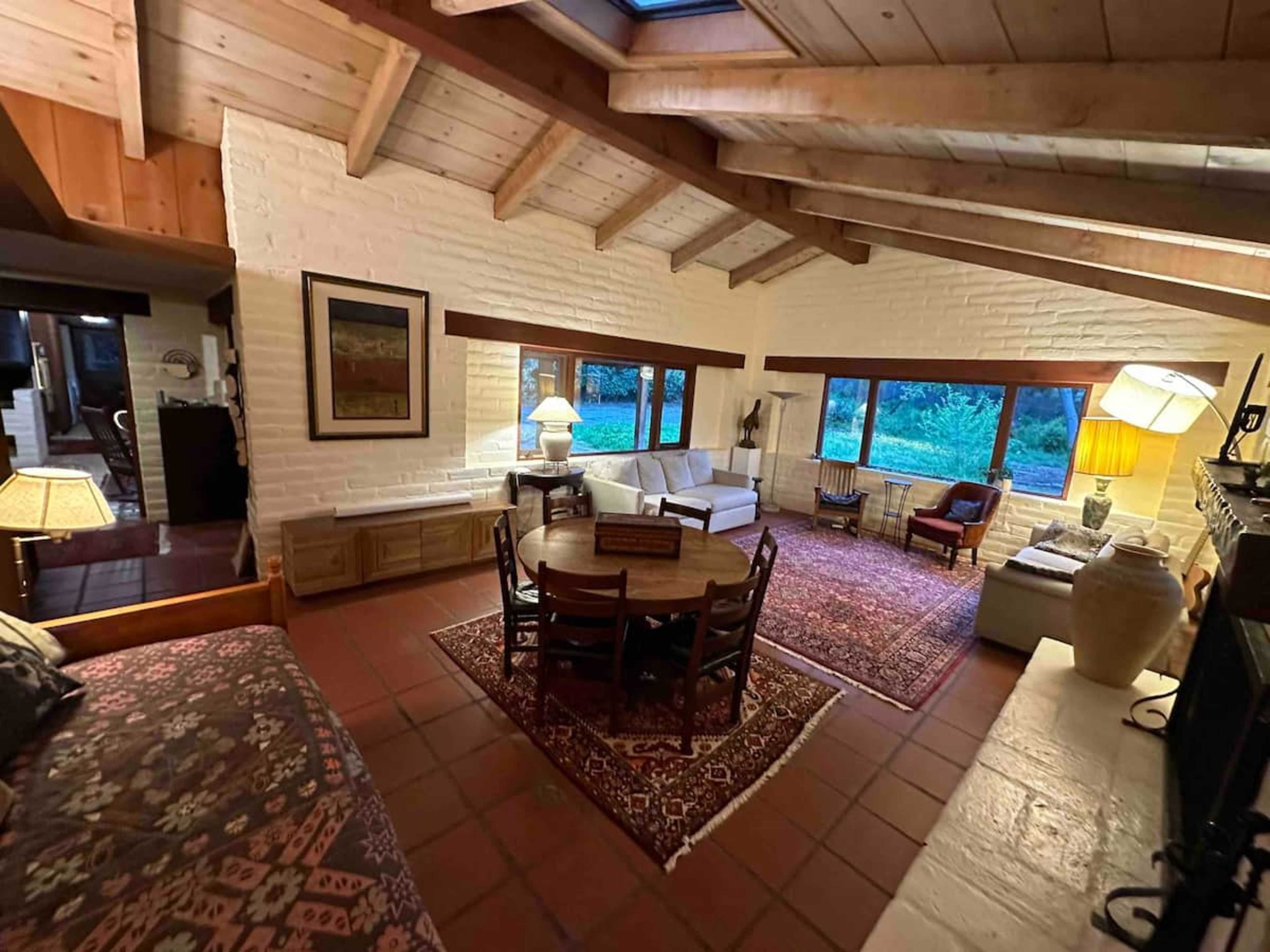 A spacious living room features a circular dining table surrounded by chairs, large windows, and wooden beams overhead.