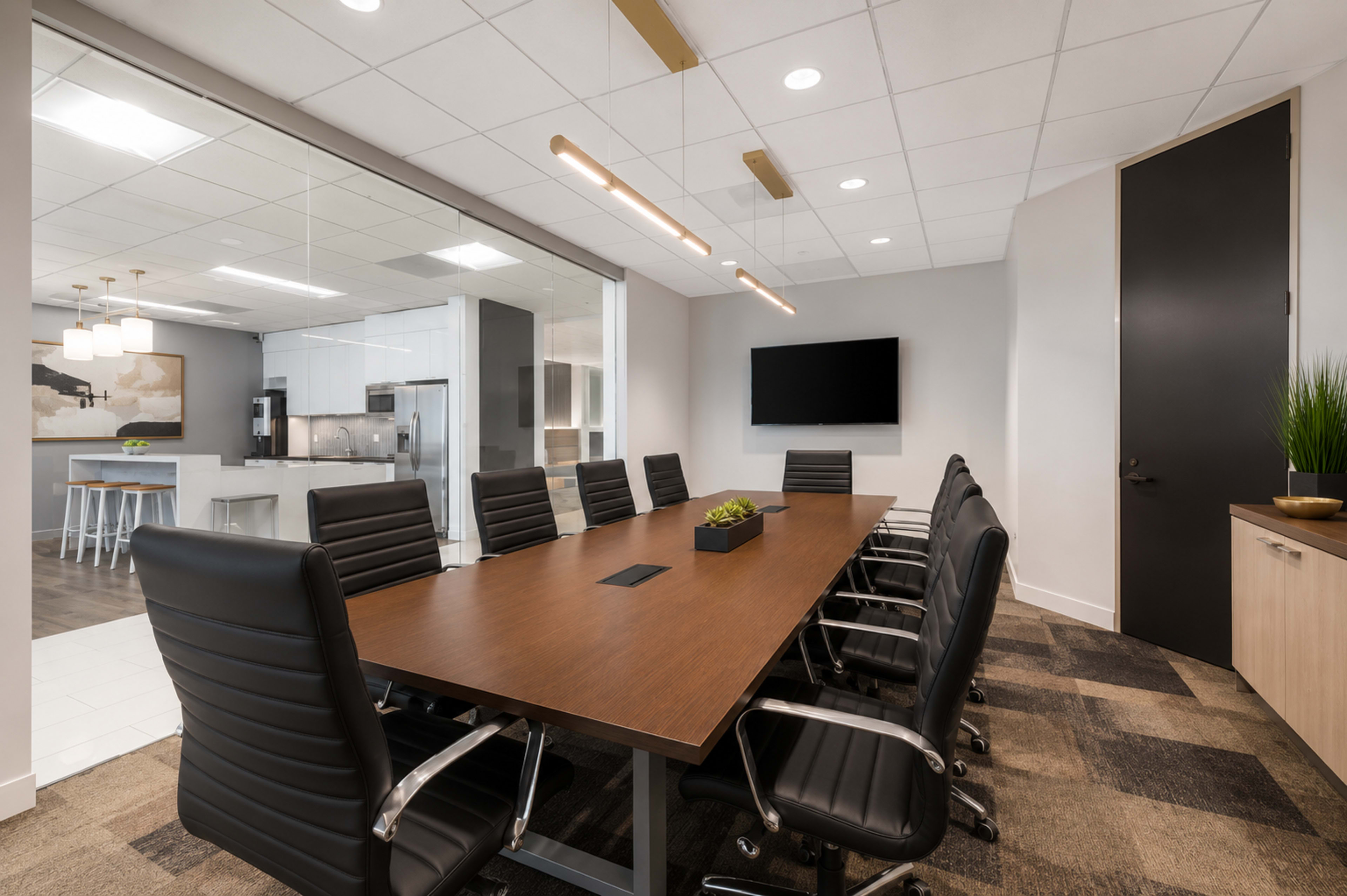 The image shows a modern conference room featuring a long wooden table surrounded by black leather chairs, with a kitchen area visible through a glass wall in the background.