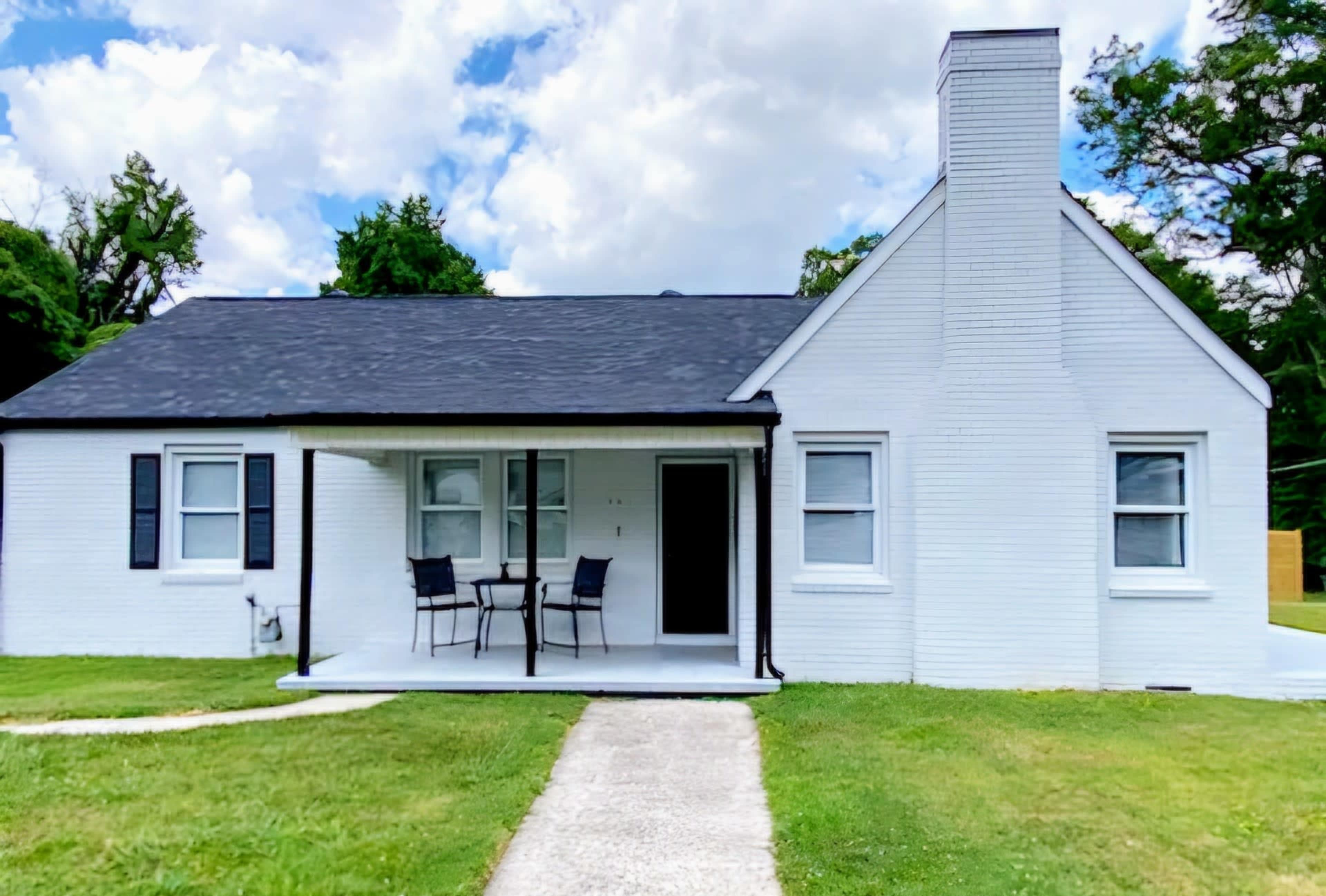 A single-story white house with a black roof features a covered porch and two chairs in front.