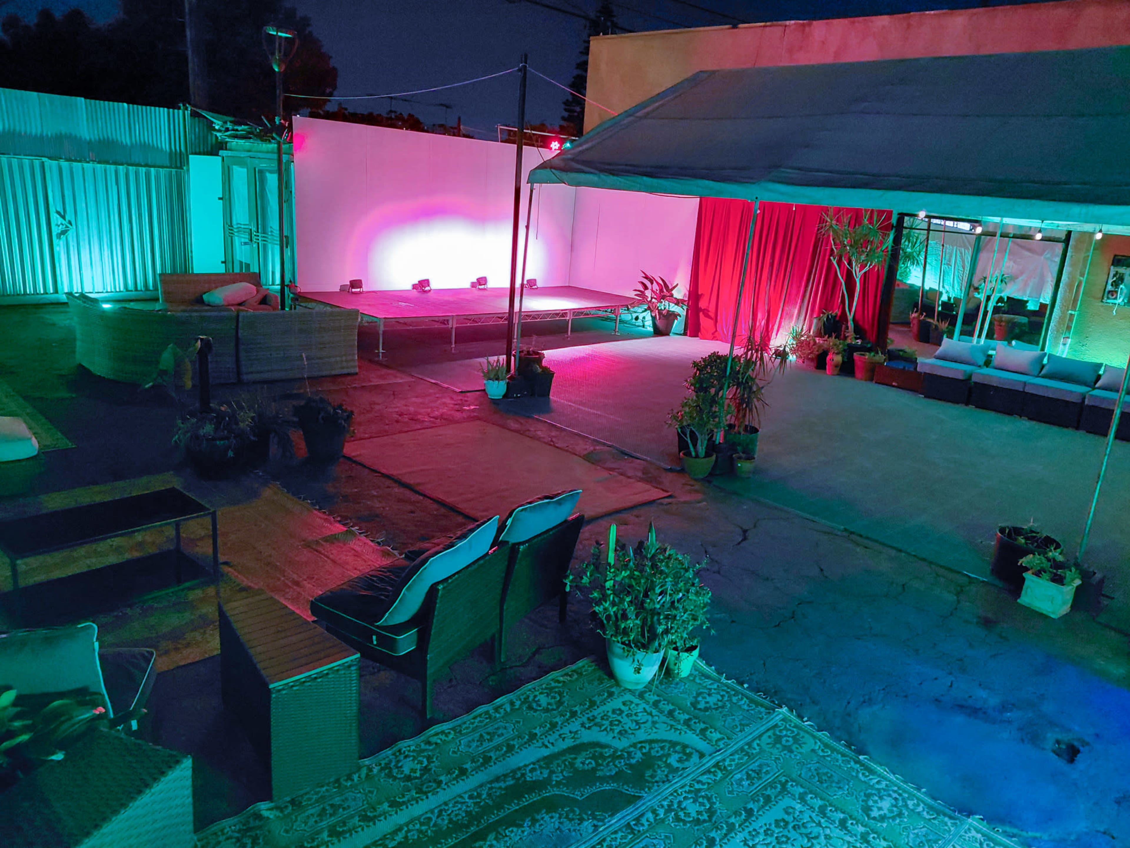 The image shows an outdoor event space with seating, a raised stage featuring spotlights, and colorful lighting creating a vibrant atmosphere.