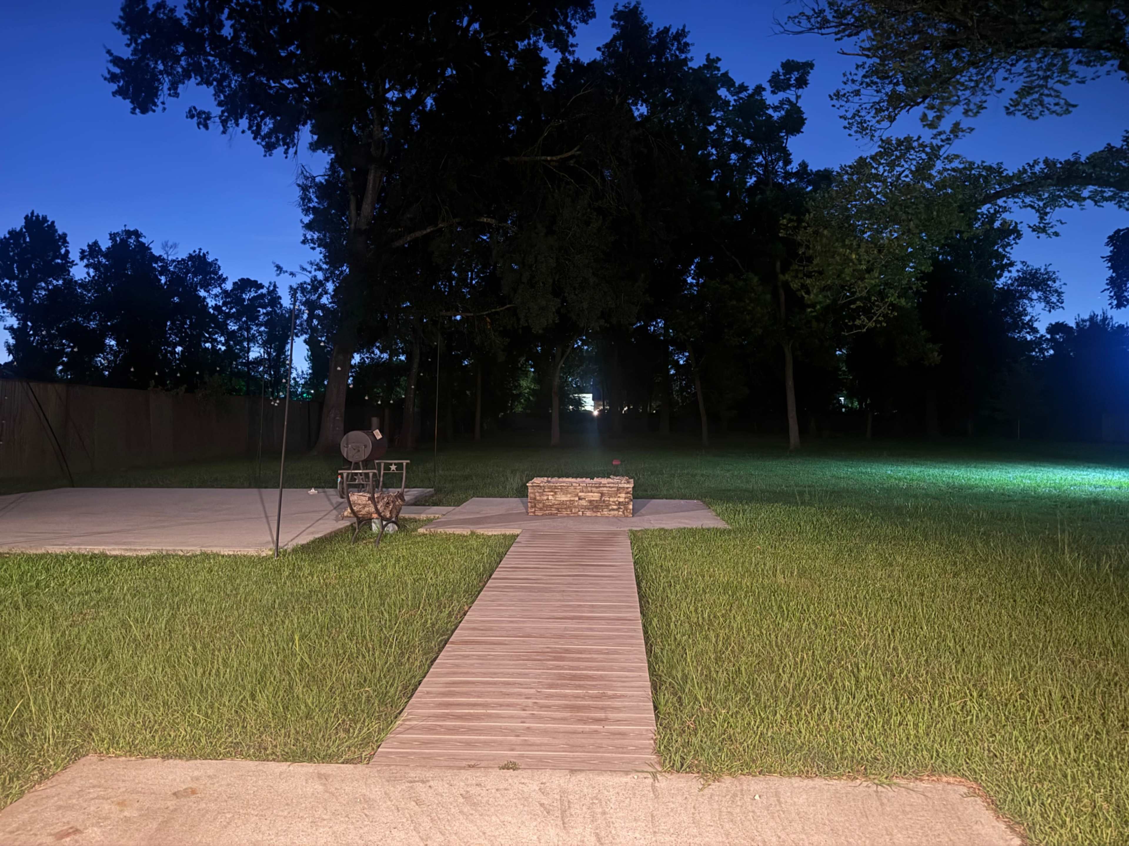The image shows a wooden pathway leading to a stone fire pit in a grassy area surrounded by trees at dusk.