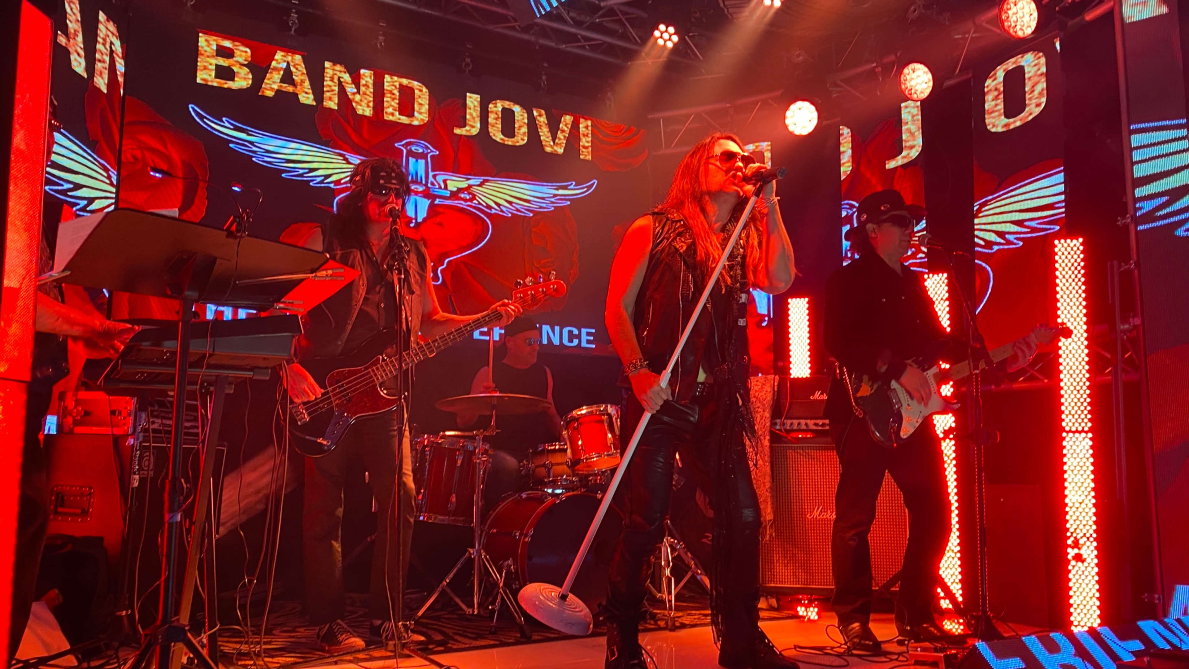 A rock band performs on stage, illuminated by vibrant red lights and large screens displaying the band’s name and emblem.