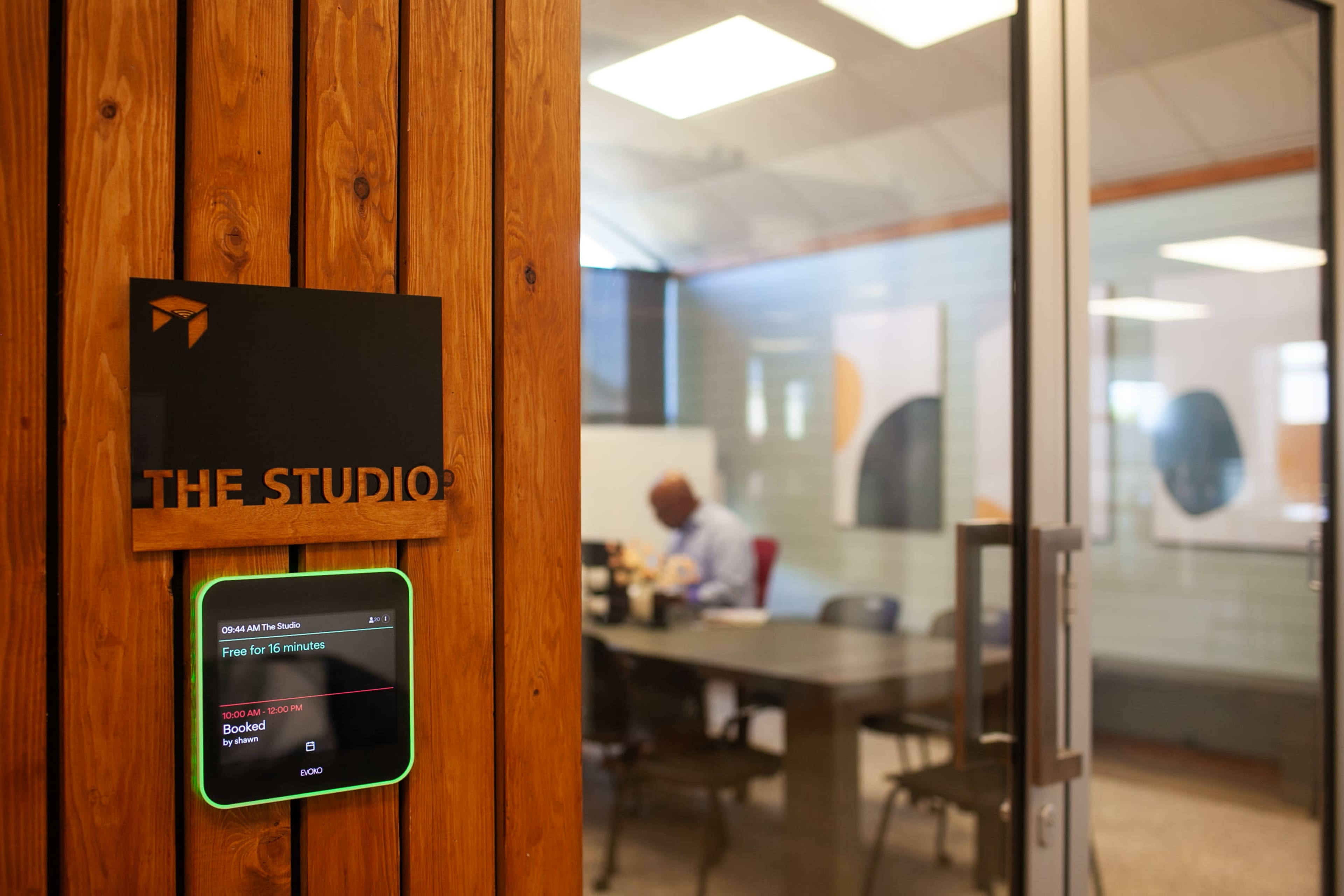 A sign for "THE STUDIO" beside a digital availability display outside a conference room with a man sitting at a table inside.