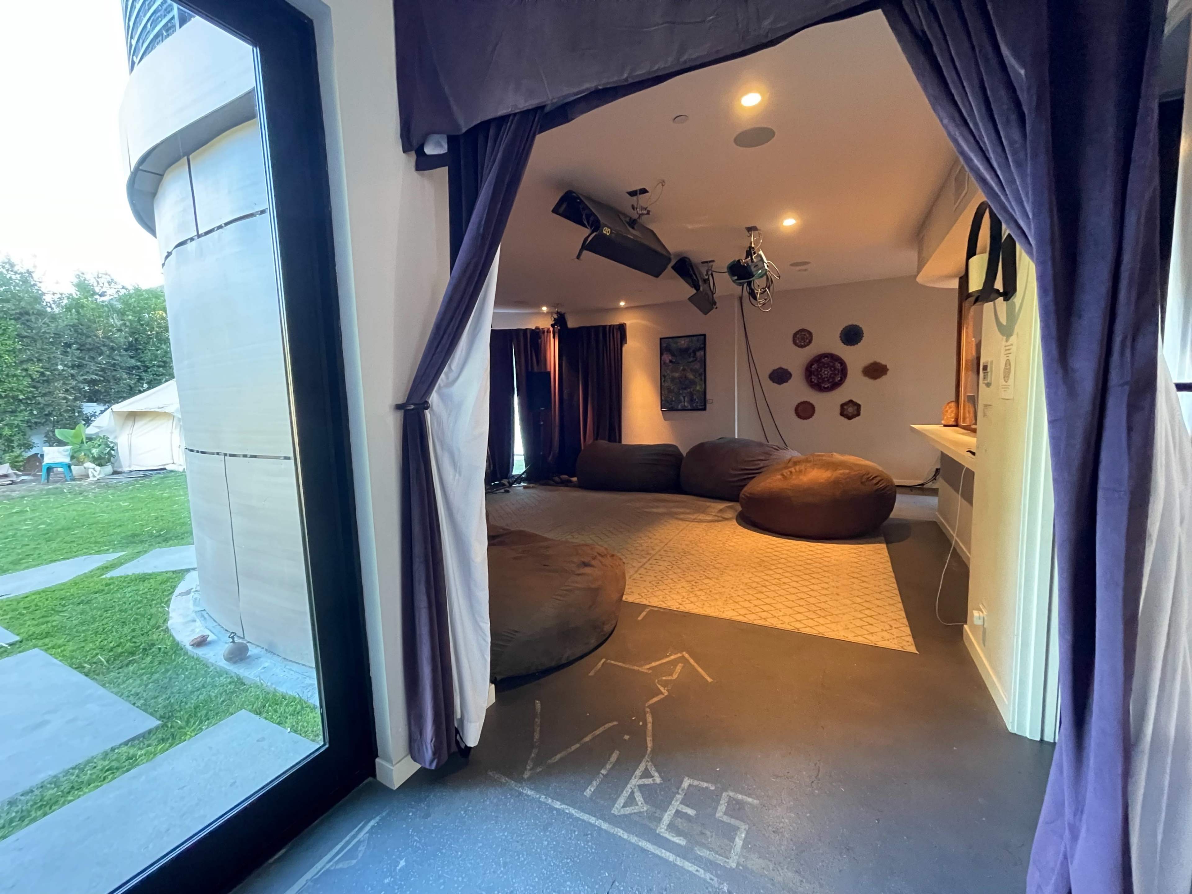 A room with large bean bags, dark curtains, and a patterned rug, leading to a grass area outside.