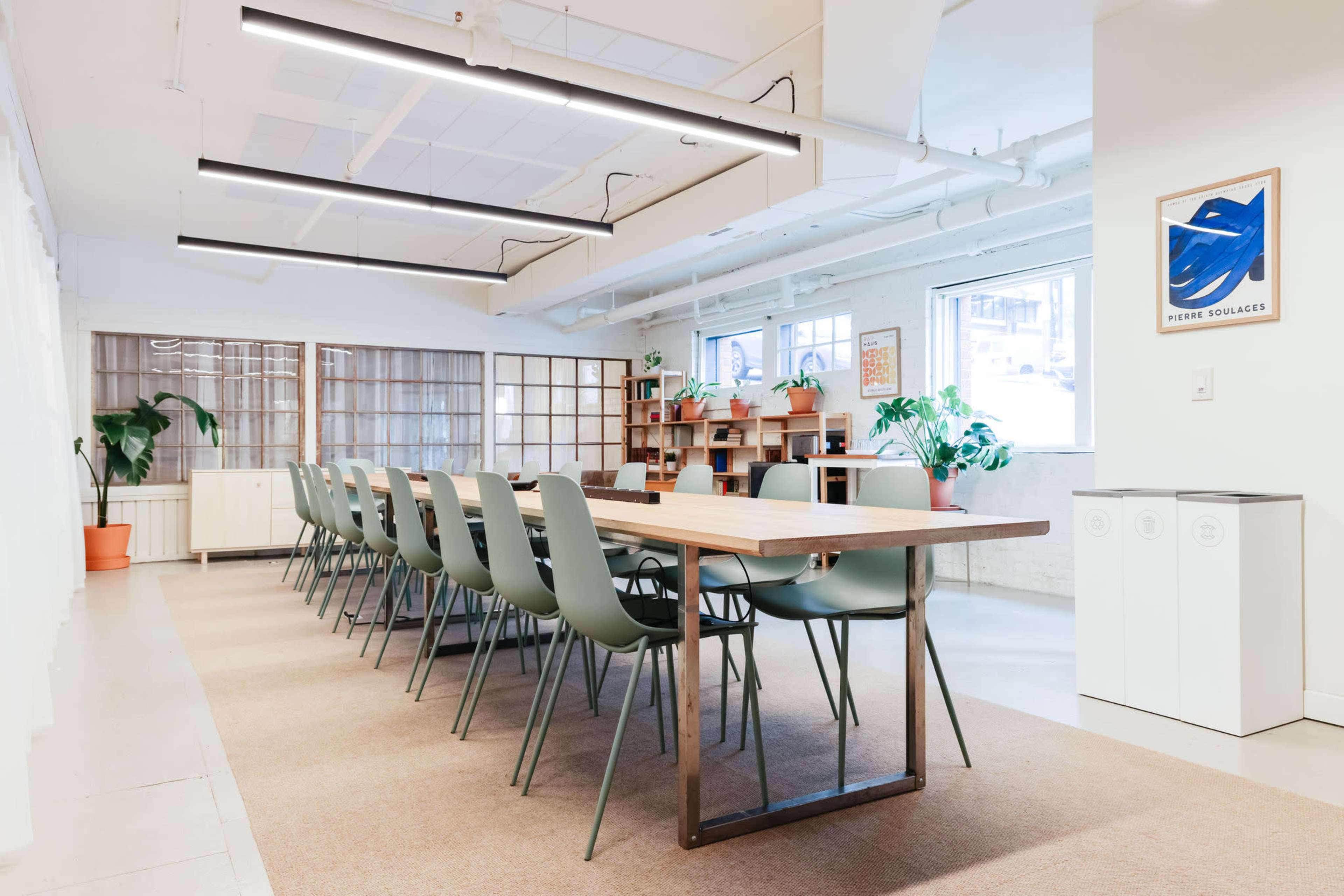A modern conference room features a long wooden table surrounded by green chairs, with large windows and decorative plants in the background.