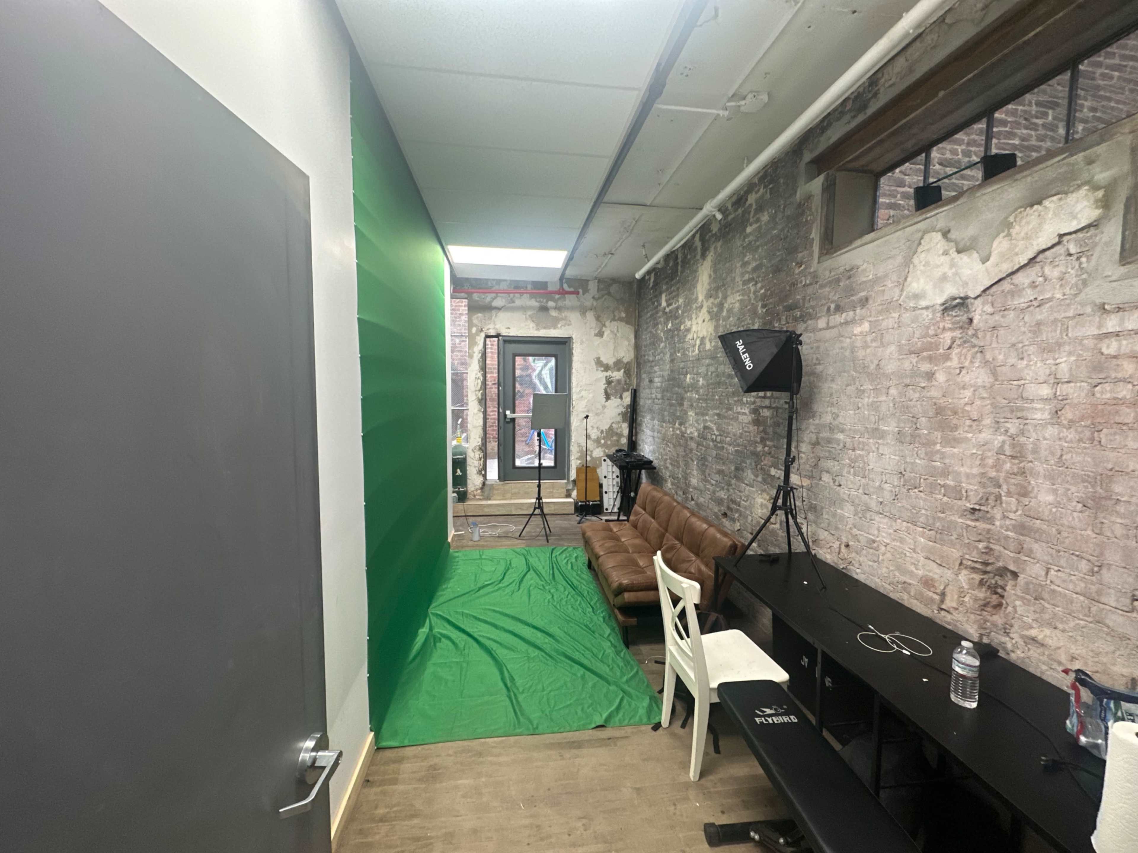 The image shows a sparse room with exposed brick walls, a green backdrop, a sofa, softbox lighting equipment, and a table against one side.