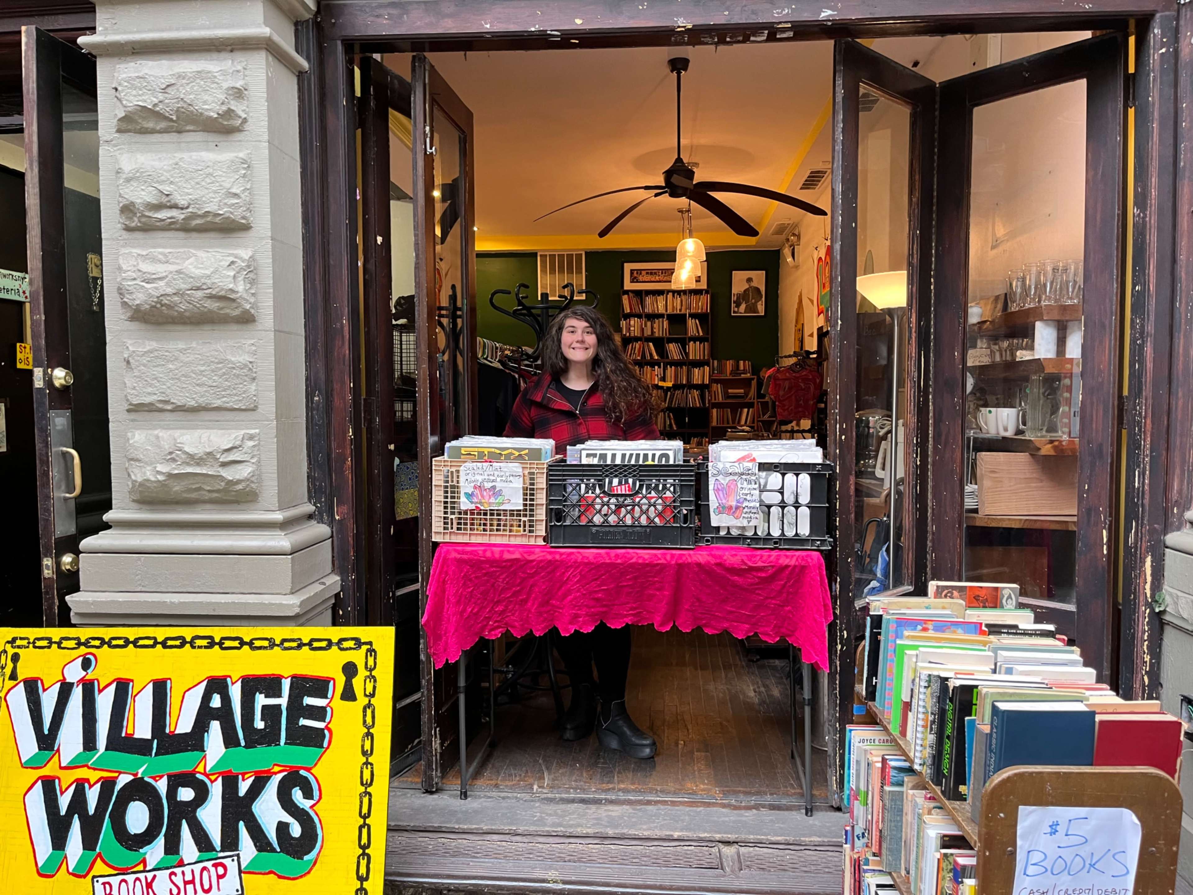 A woman stands behind a display table at the entrance of a bookstore filled with books and various items.