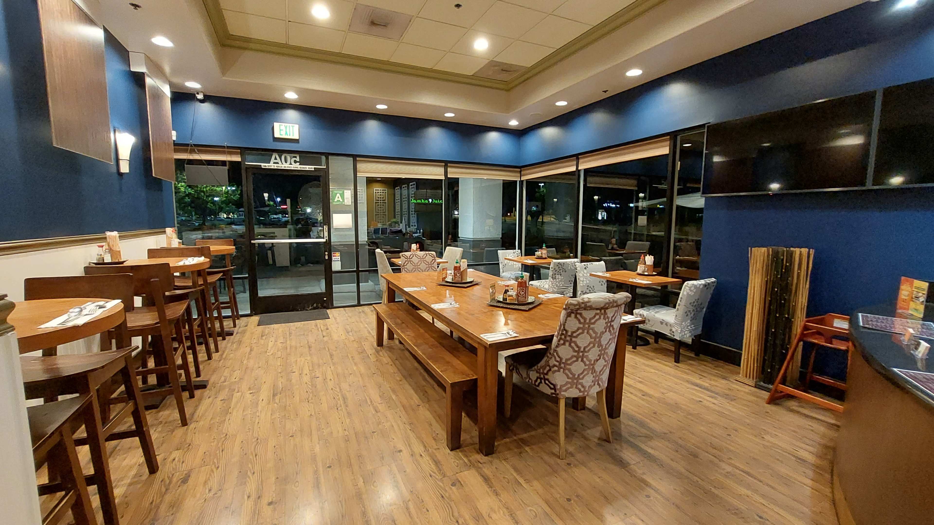 The image shows a dining area of a restaurant featuring wooden tables and chairs, upholstered seating, and large windows illuminating the space.