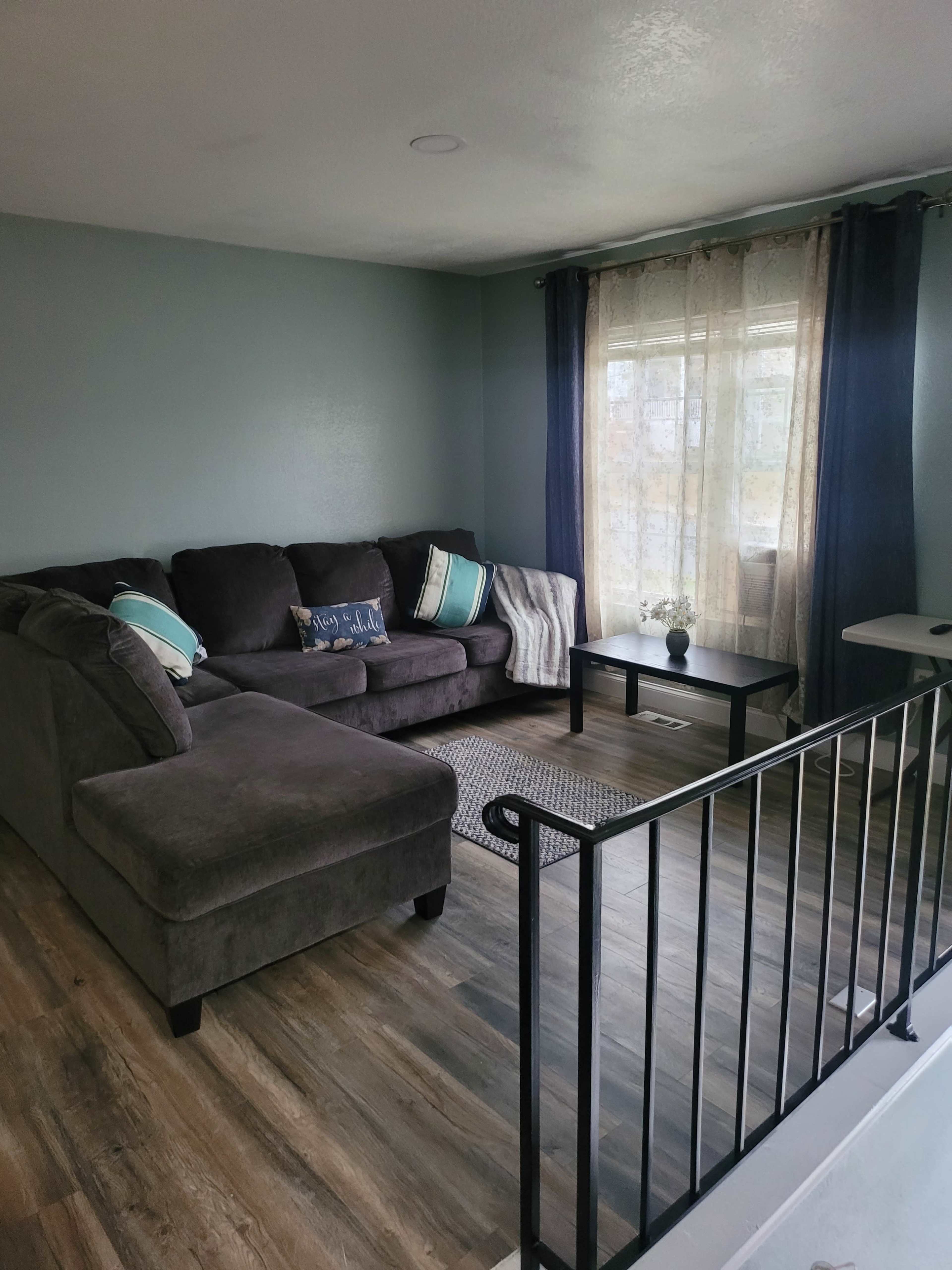 The image shows a living room with a gray sectional sofa, a small black coffee table, and a window dressed with sheer and dark curtains.