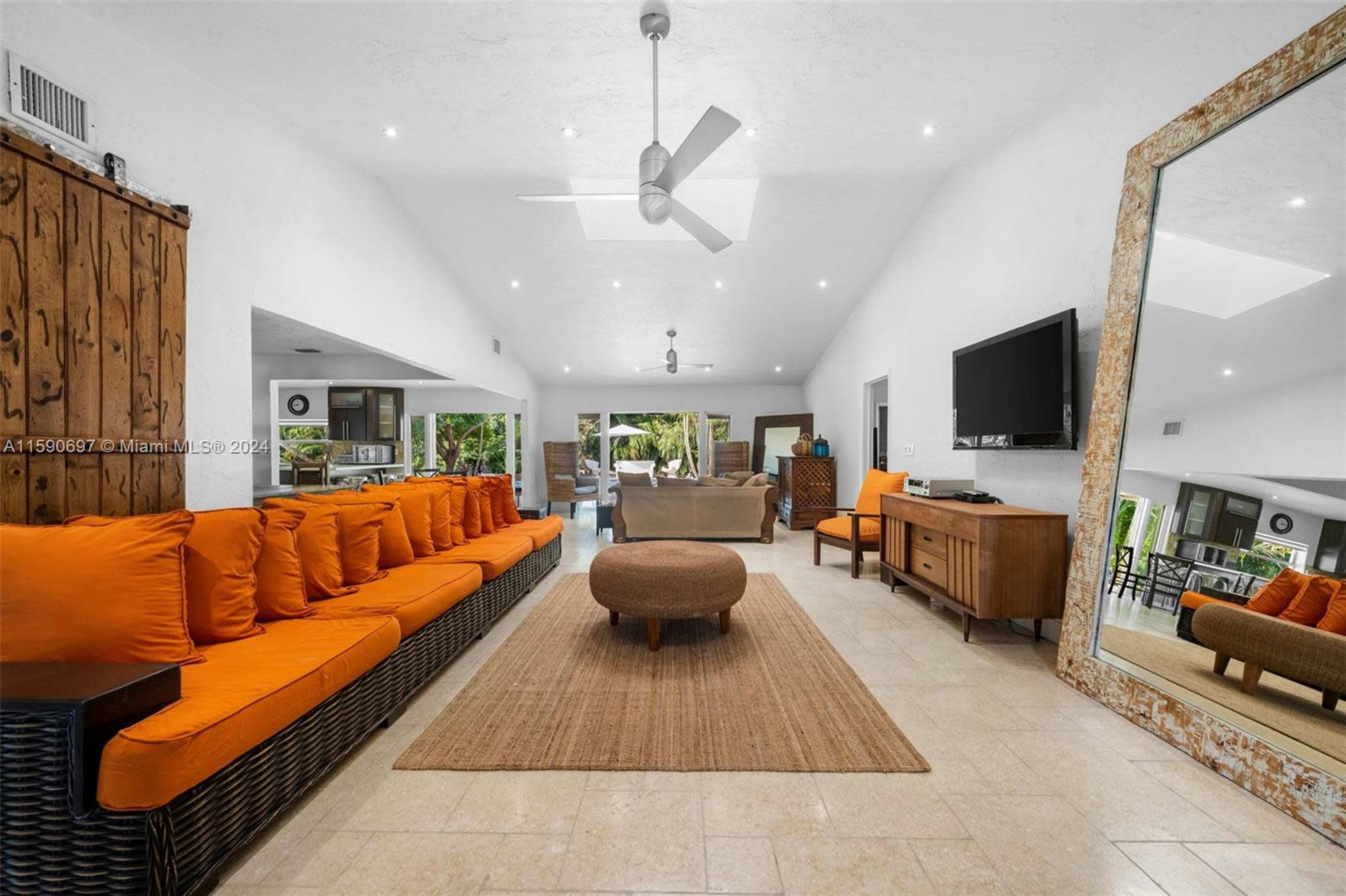 The image shows a spacious interior living area featuring a large orange-cushioned sofa, a central round coffee table, a flat-screen TV on the wall, and a mirror reflecting part of the room.