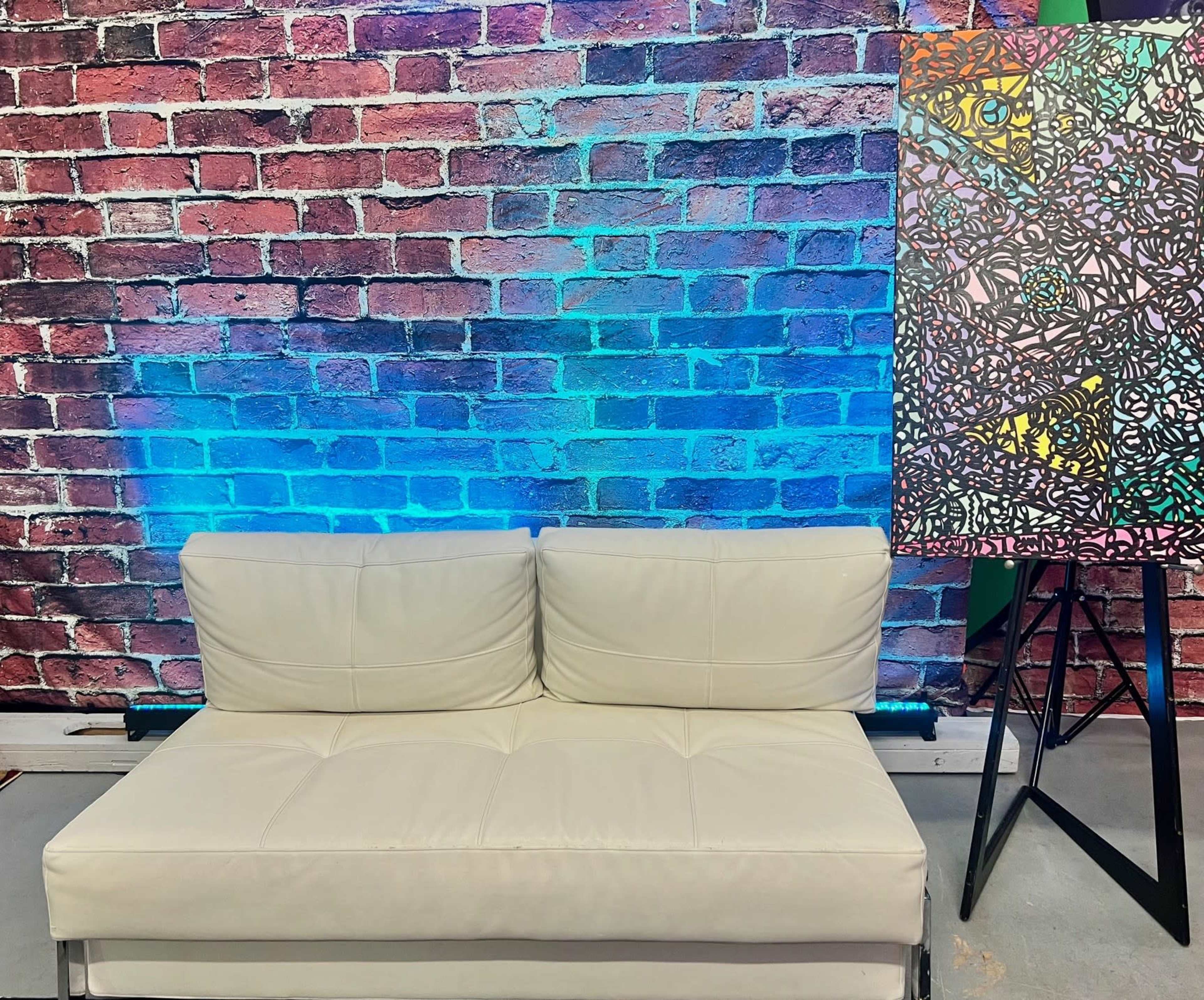 The scene features a white couch in front of a colorful brick wall and a vibrant artwork displayed on an easel.
