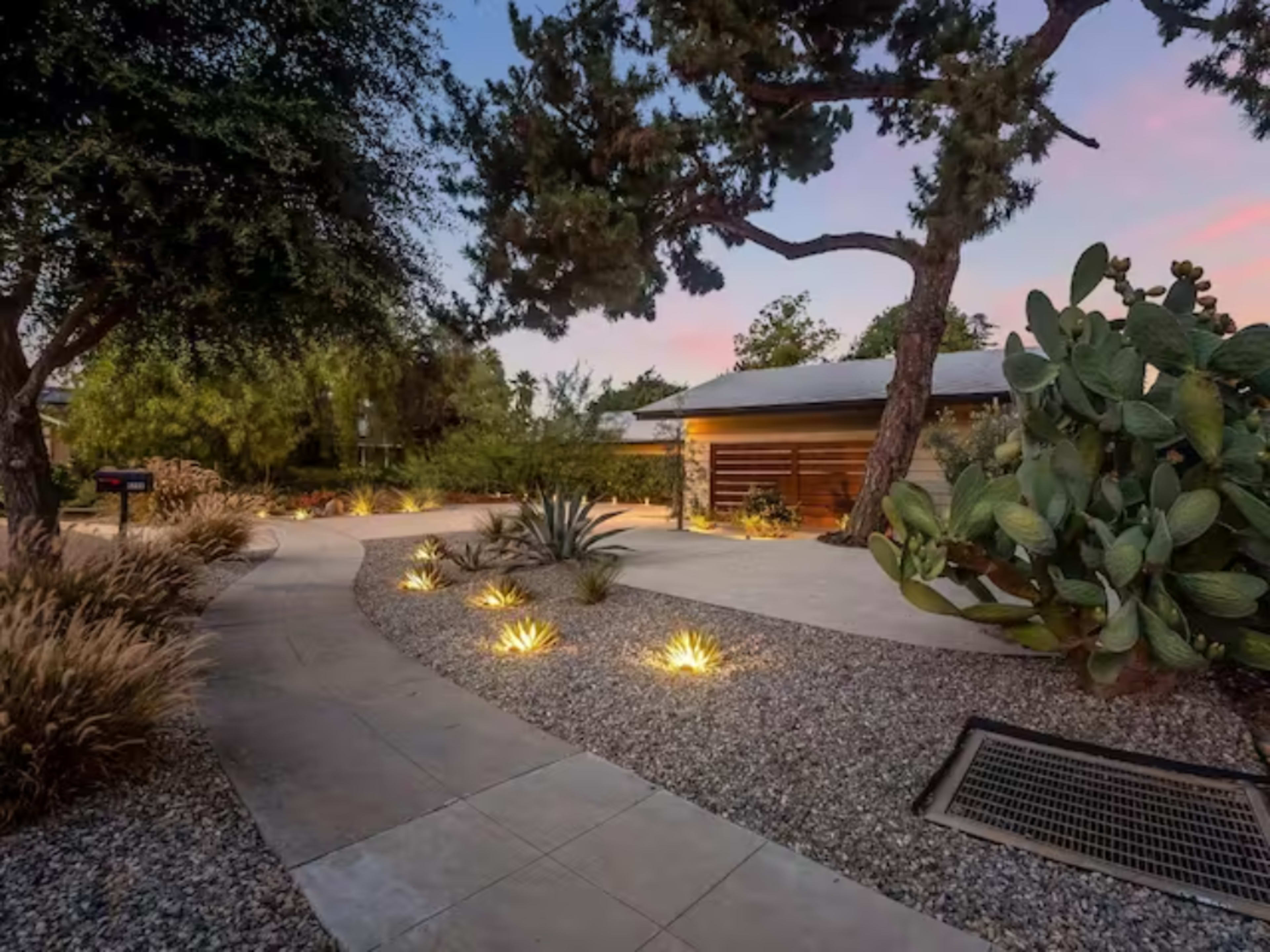 A winding pathway lined with gravel and plants leads to a modern home set among trees and desert vegetation.