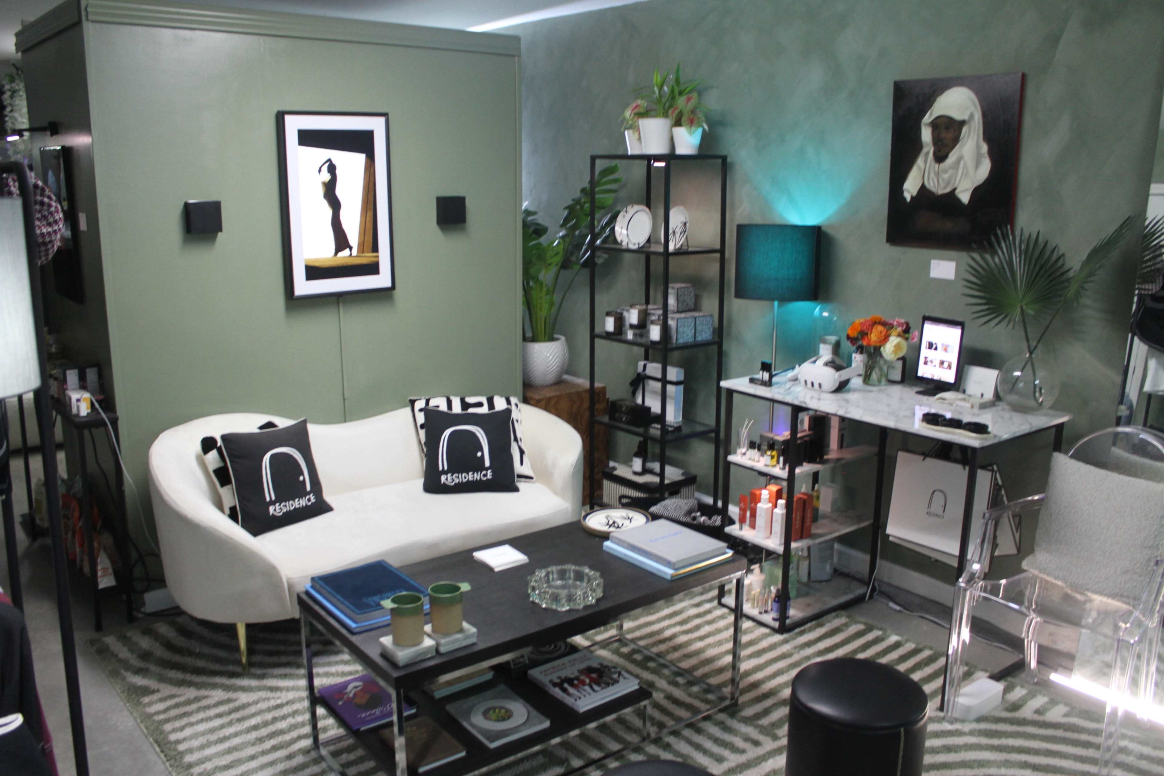 The image shows a stylish living room with a curved white sofa, a coffee table with various items on it, and a decorative shelf with plants and art pieces against a green wall.