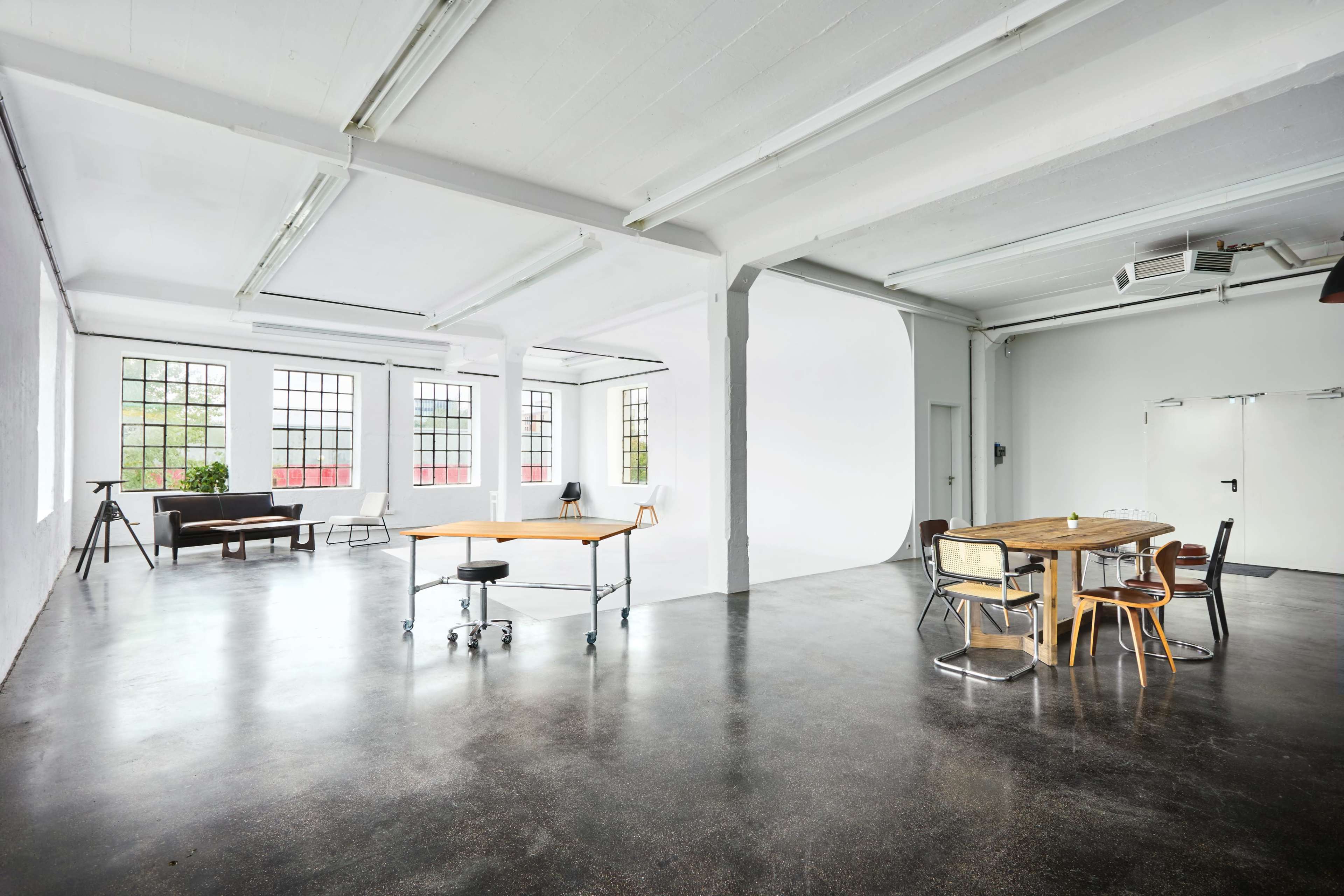 The image shows a spacious, bright studio or office area with a large table on wheels and various seating options, complemented by large windows allowing natural light.