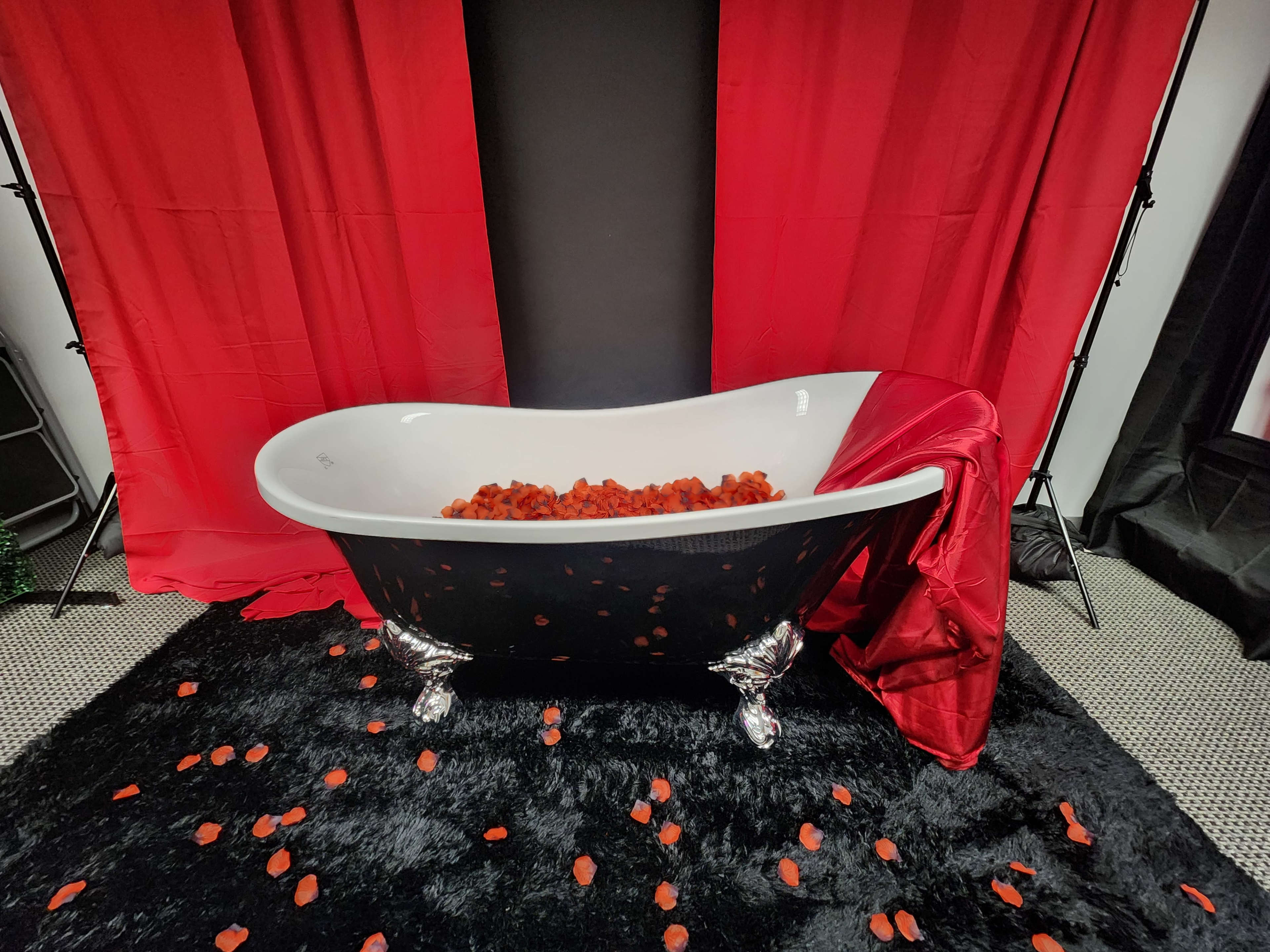 A black and white freestanding bathtub filled with red rose petals, surrounded by red and black fabric backdrops.