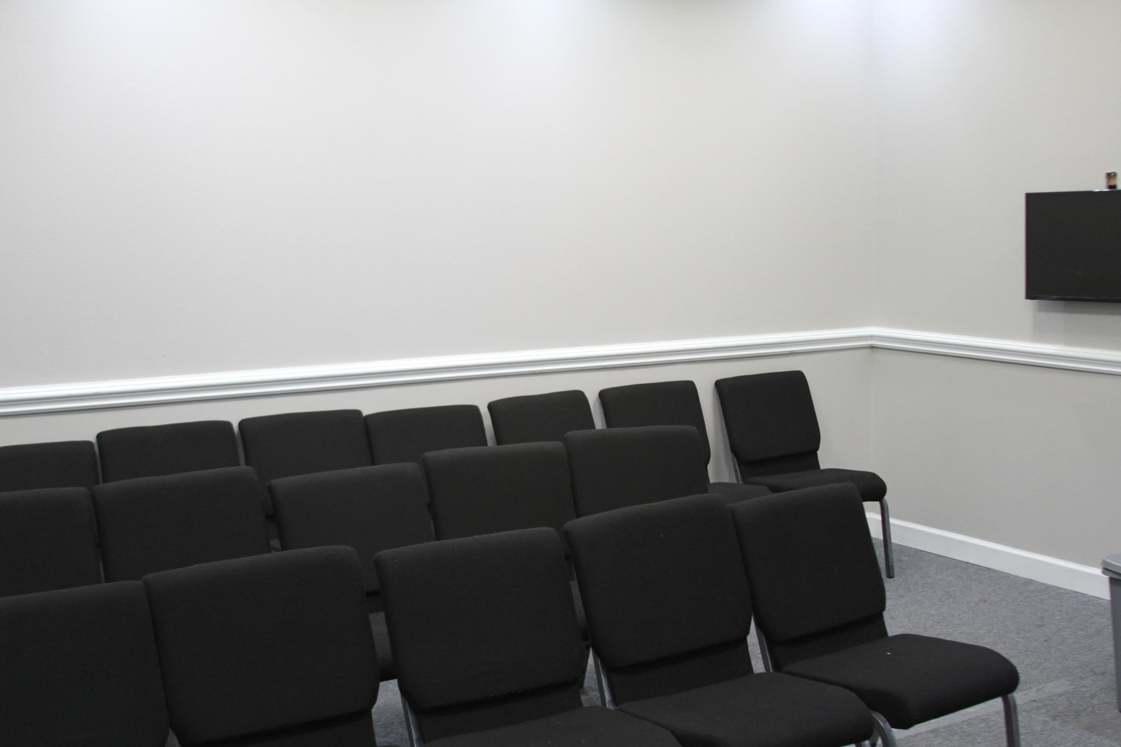 The image shows a small meeting room with rows of black chairs facing a wall-mounted television.