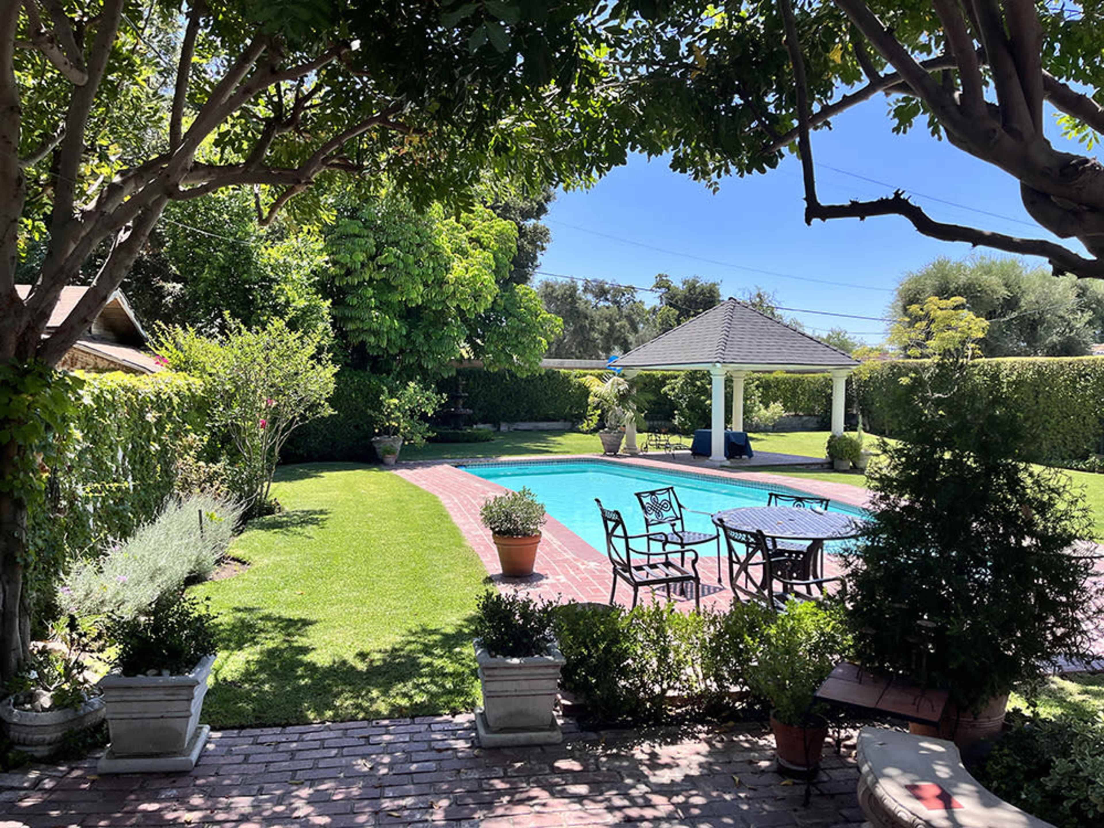 A well-maintained swimming pool is surrounded by landscaped greenery and patio furniture in a sunny backyard.