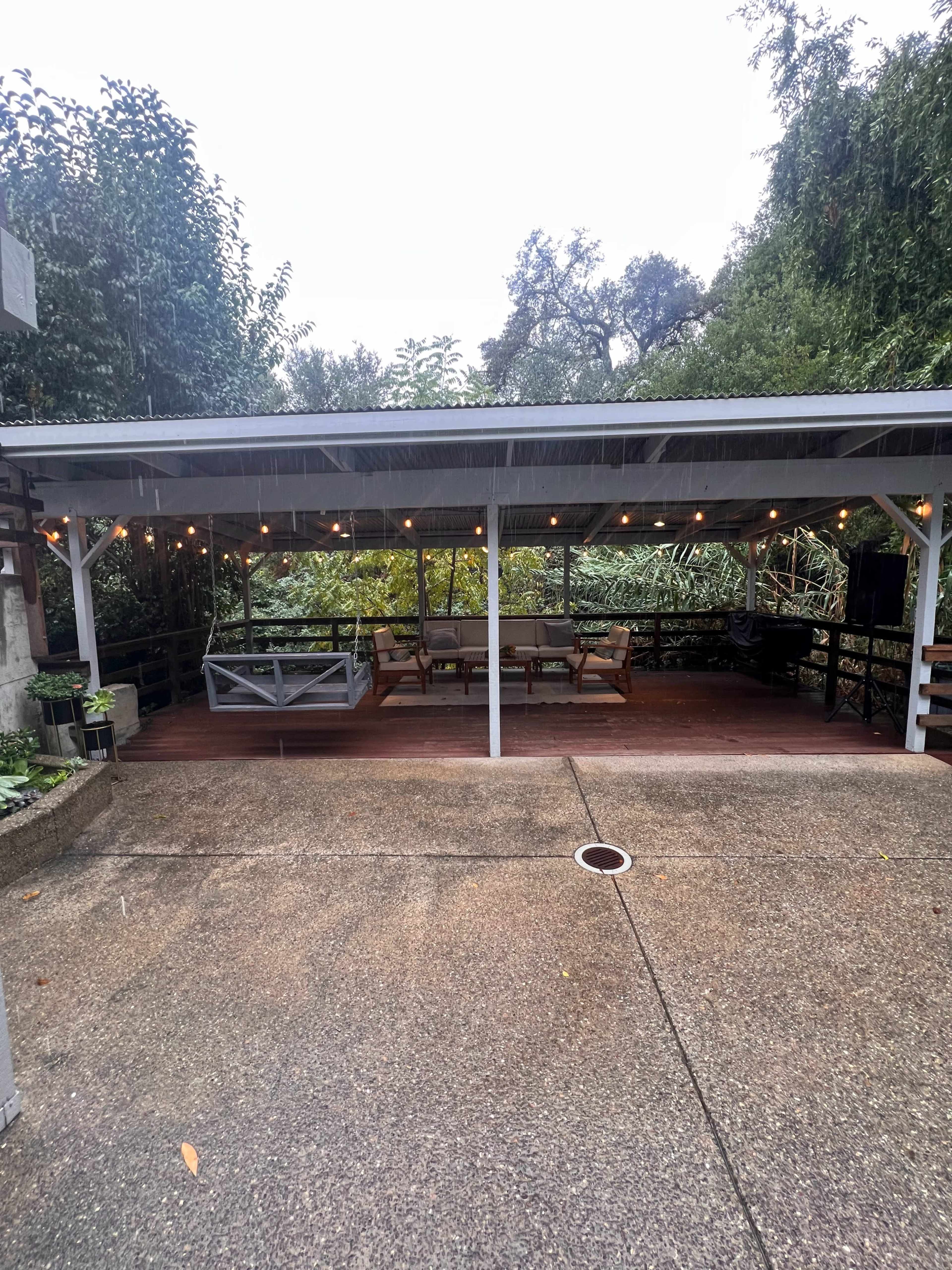 The image shows a covered outdoor patio area with wooden furniture, surrounded by greenery and illuminated by string lights.