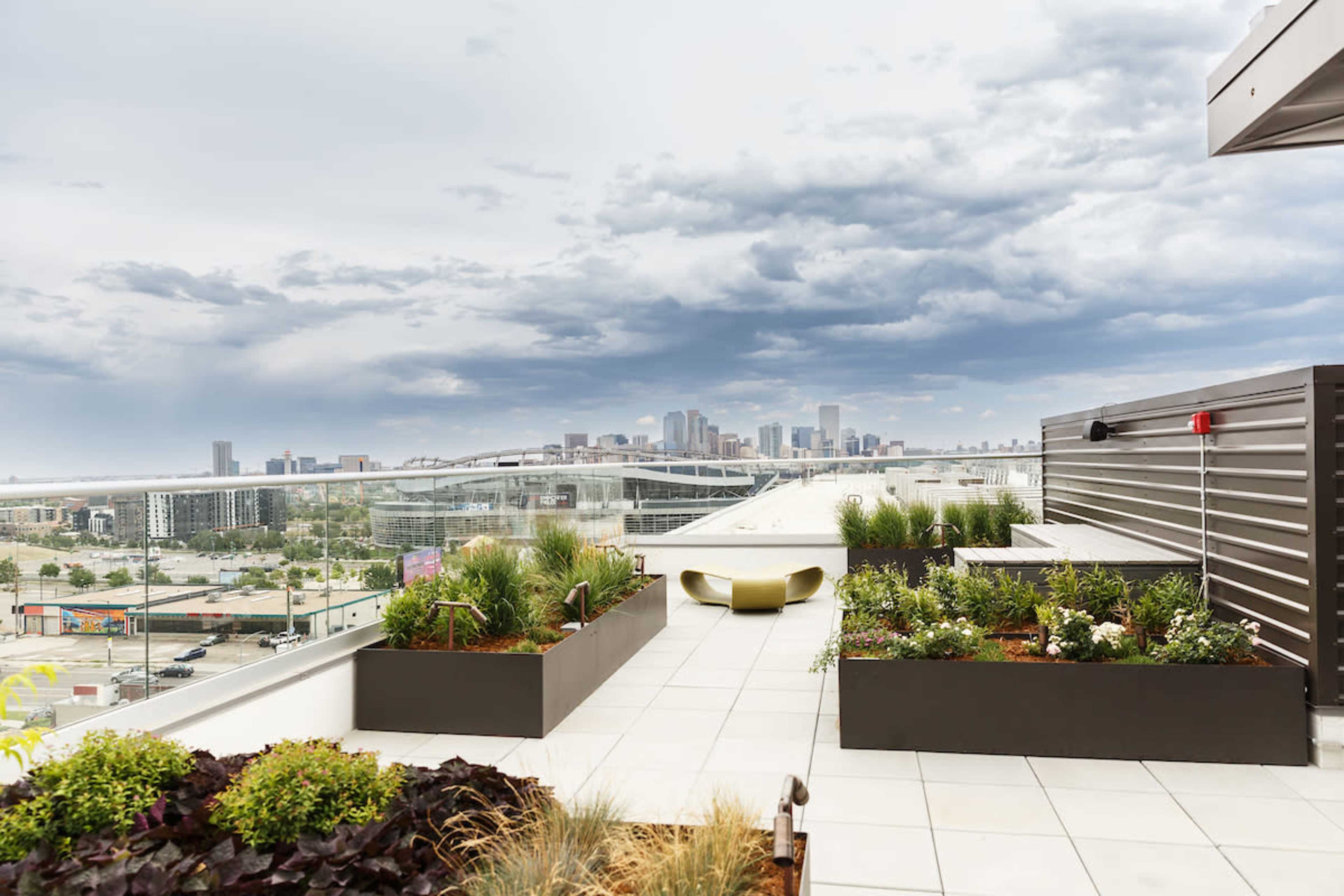 The image shows a rooftop terrace with planter boxes, featuring a view of a city skyline under a cloudy sky.