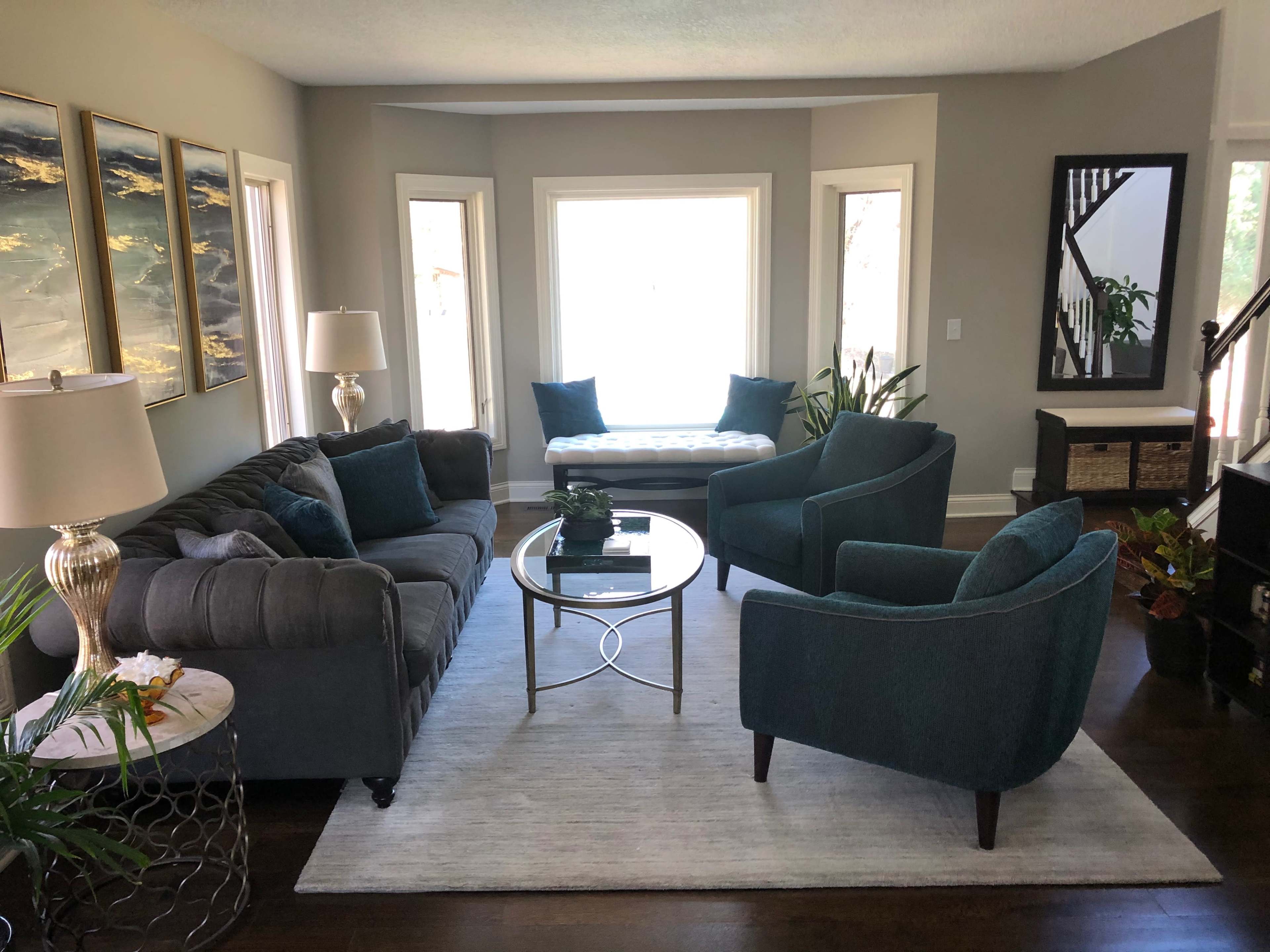 The living room features a gray sectional sofa, two teal armchairs, a round glass coffee table, and decorative artwork on the walls.
