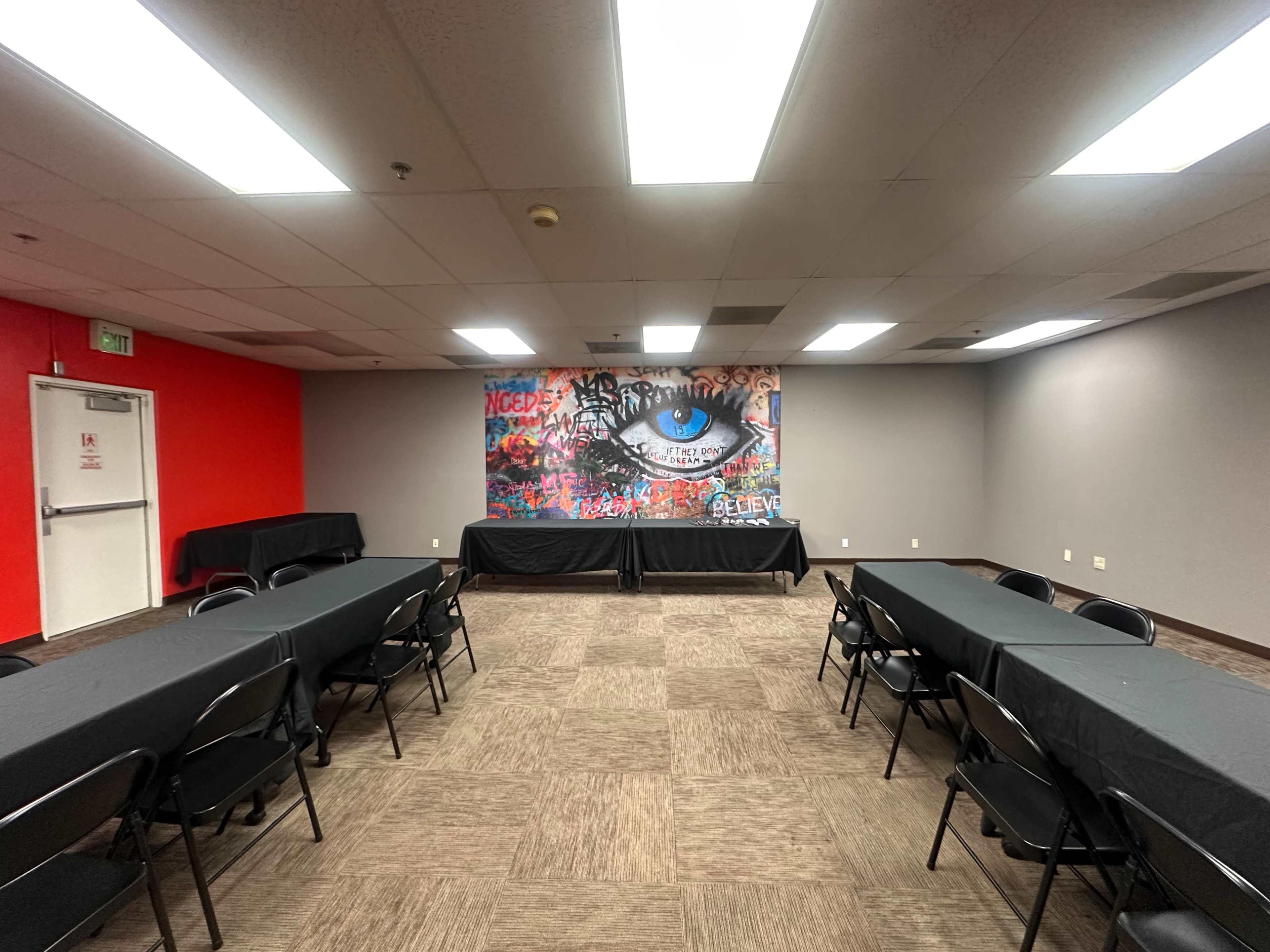 The image shows an empty meeting room with multiple rows of black tables and a colorful mural of an eye on the wall.