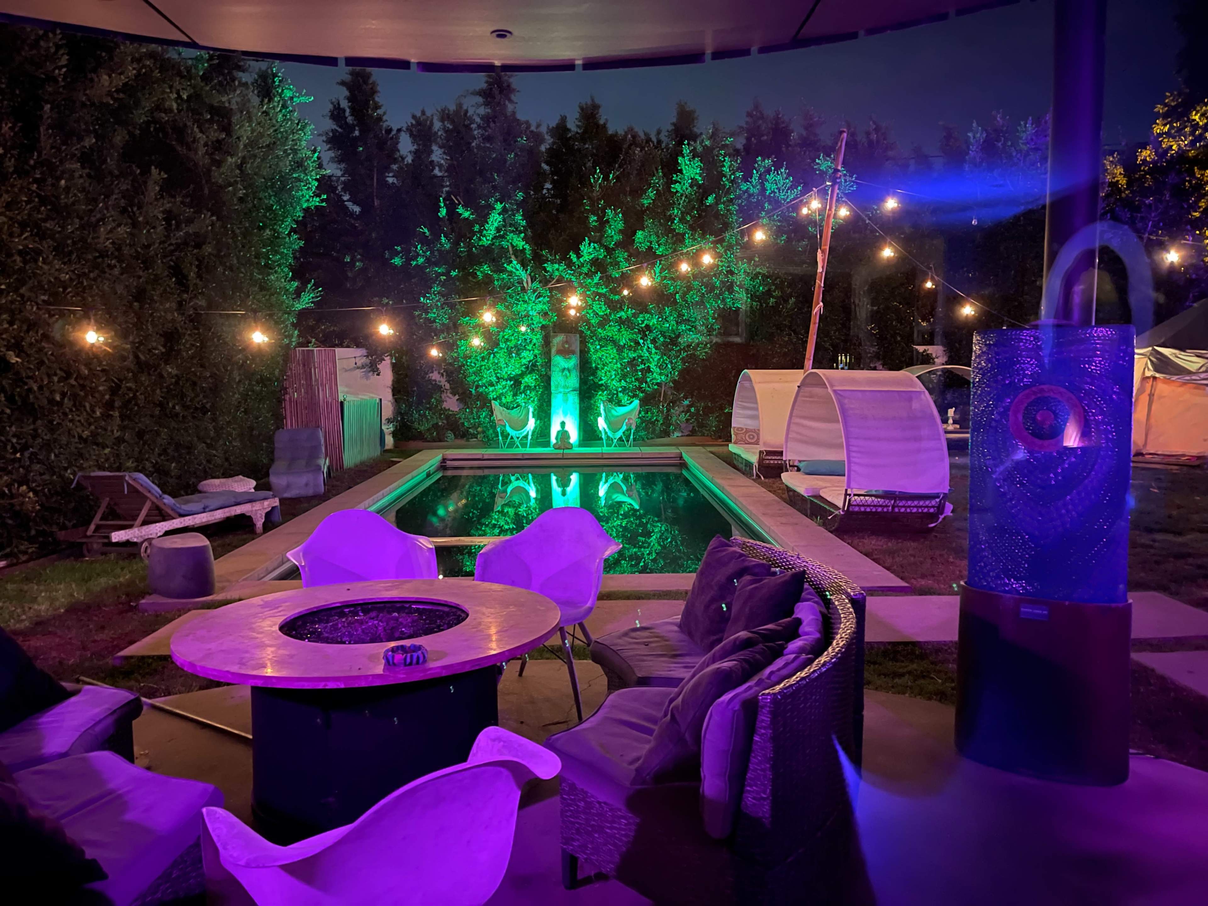 A pool area at night, featuring a circular fire pit surrounded by seating, illuminated by colorful lights and string lights in the trees.