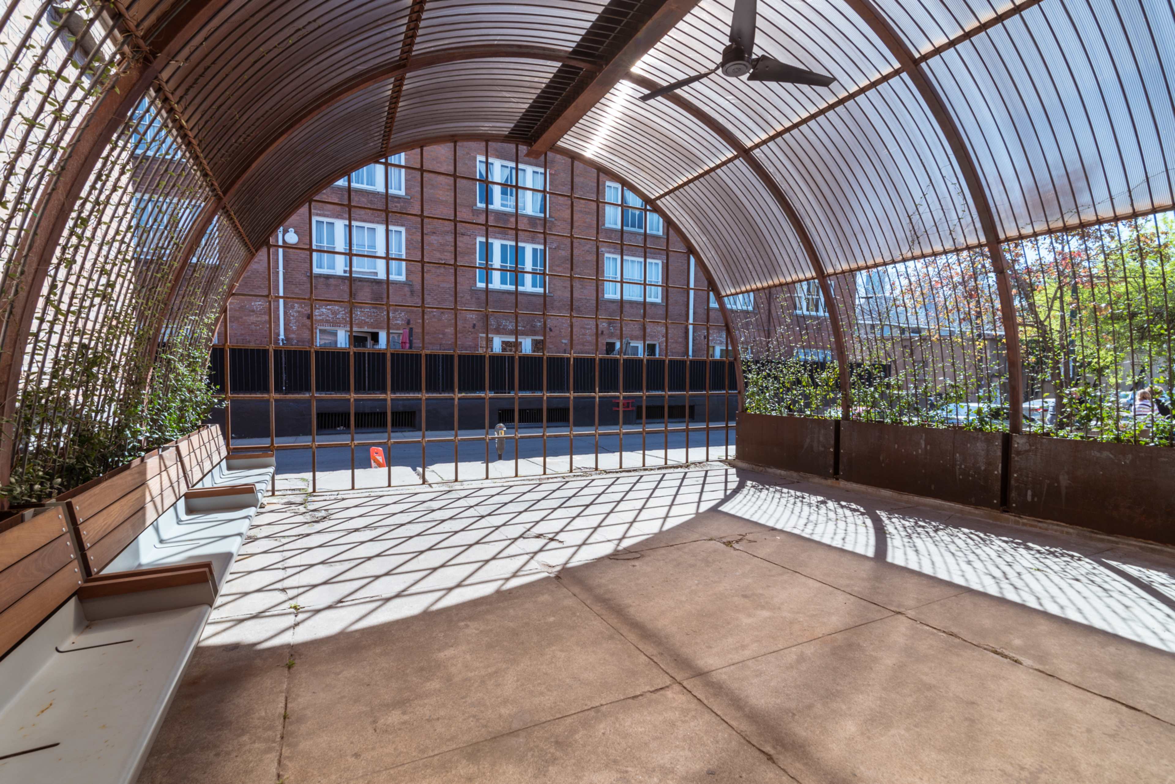 An arched structure with metal framing and wooden benches overlooks a street, casting shadows on the concrete floor.