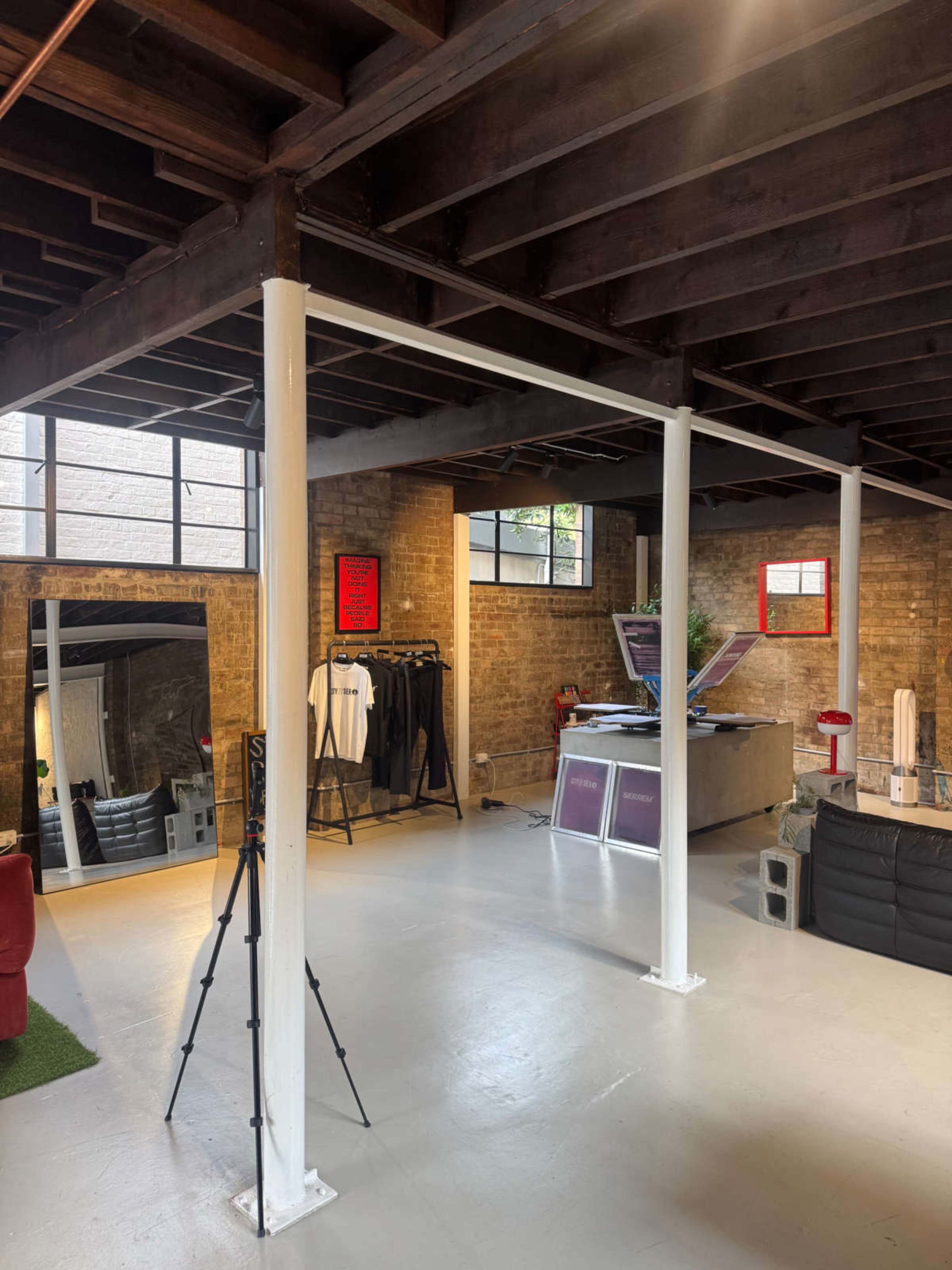 The image shows a spacious industrial-style interior with exposed brick walls, wooden beams, and various furnishings, including a clothing rack and a camera tripod.
