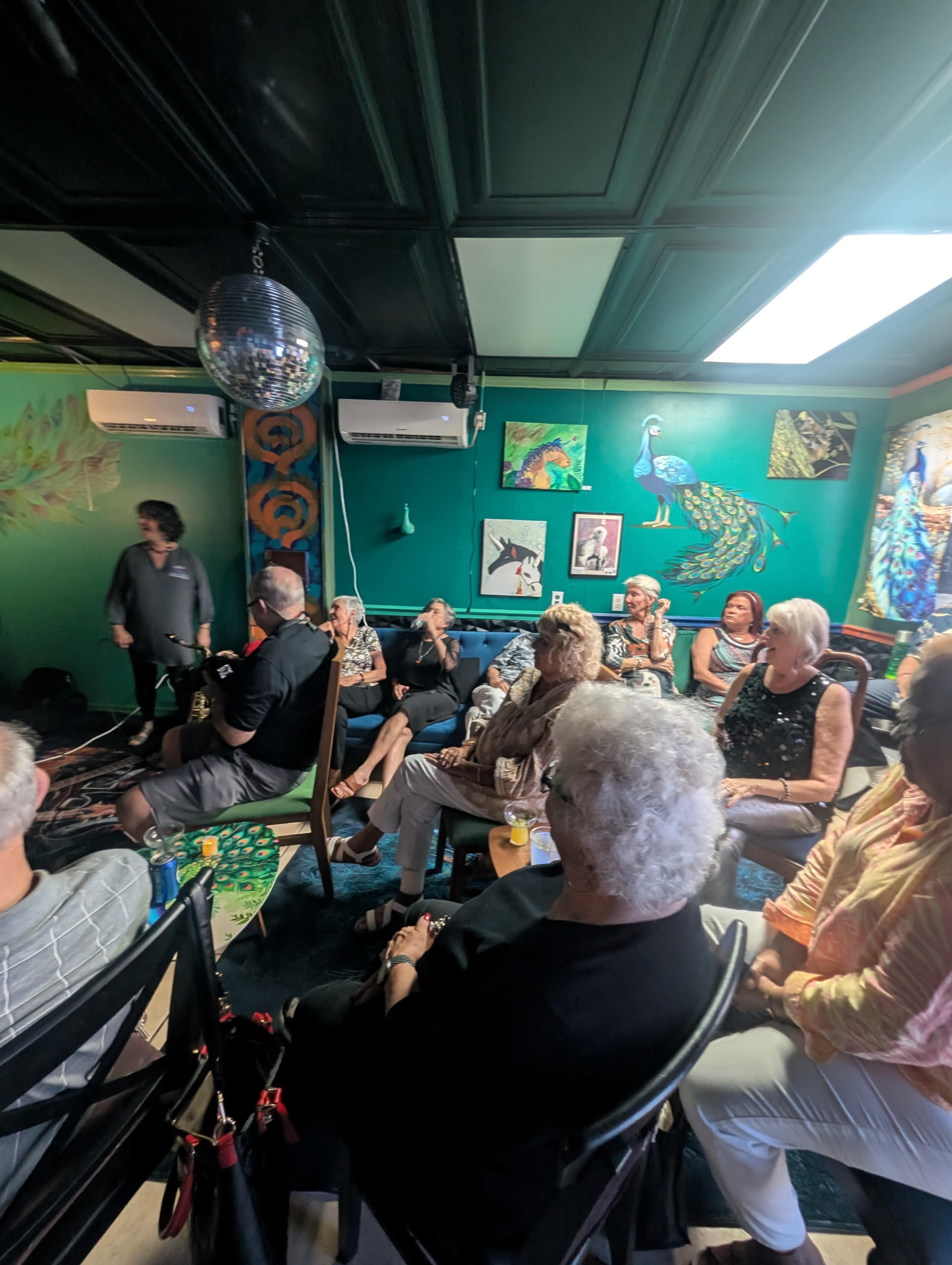 A group of people sit in a colorful room adorned with artwork, watching a performer on a small stage.