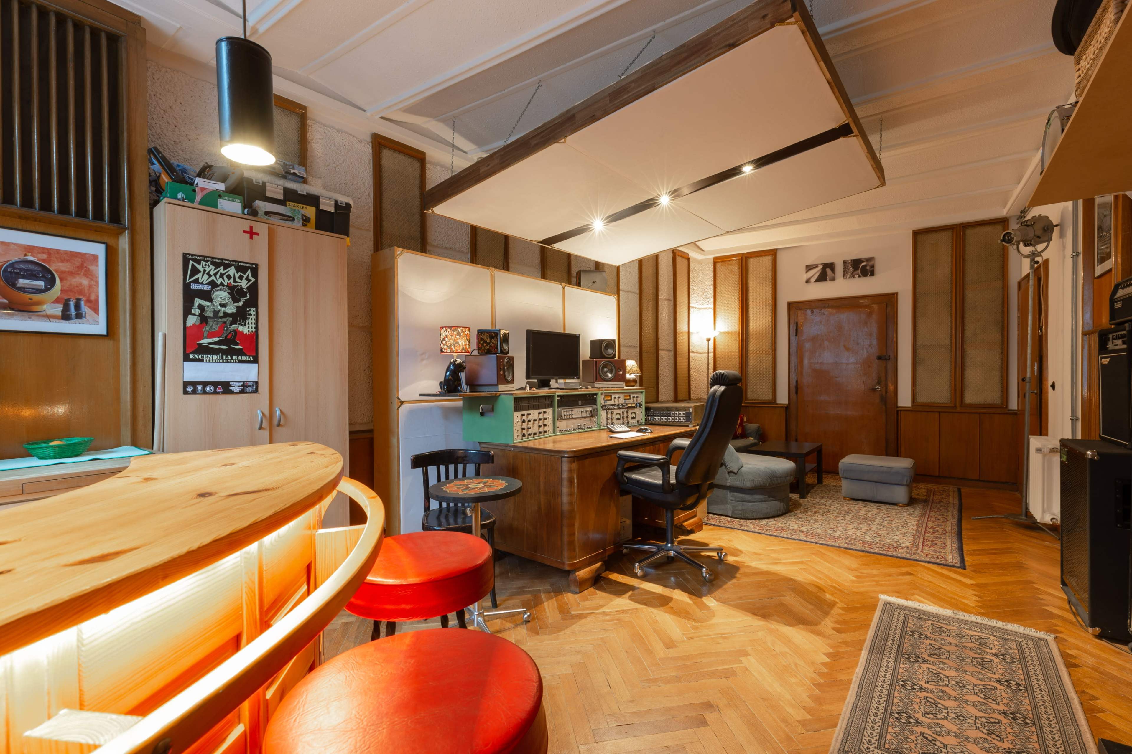 A cozy recording studio with wood paneling, a mixing desk, a comfortable chair, and a bar area with red stools.