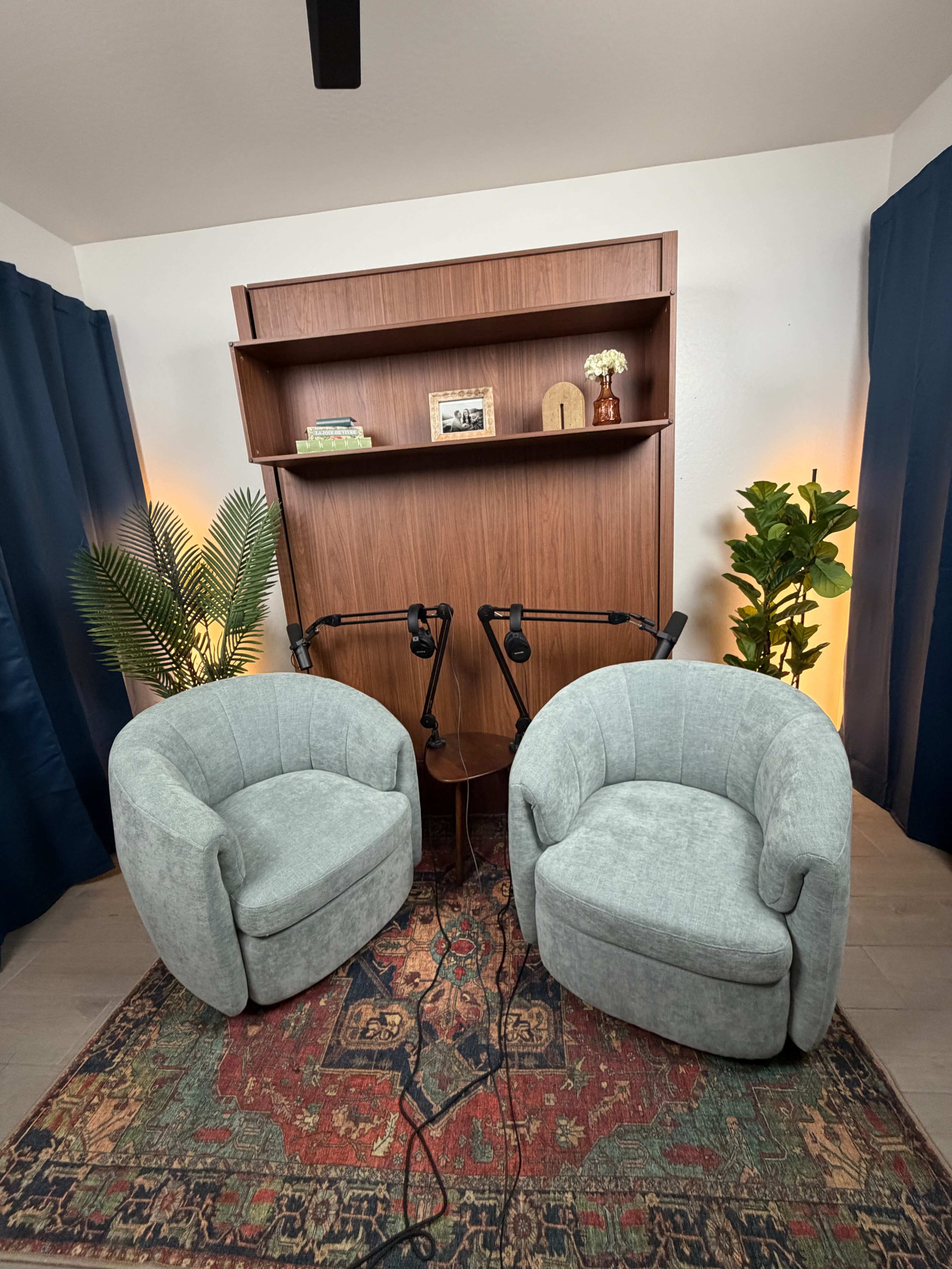 The image shows a cozy recording studio setup with two light gray chairs facing each other, a wooden shelving unit in the background, and two microphones positioned on stands in front of the chairs.