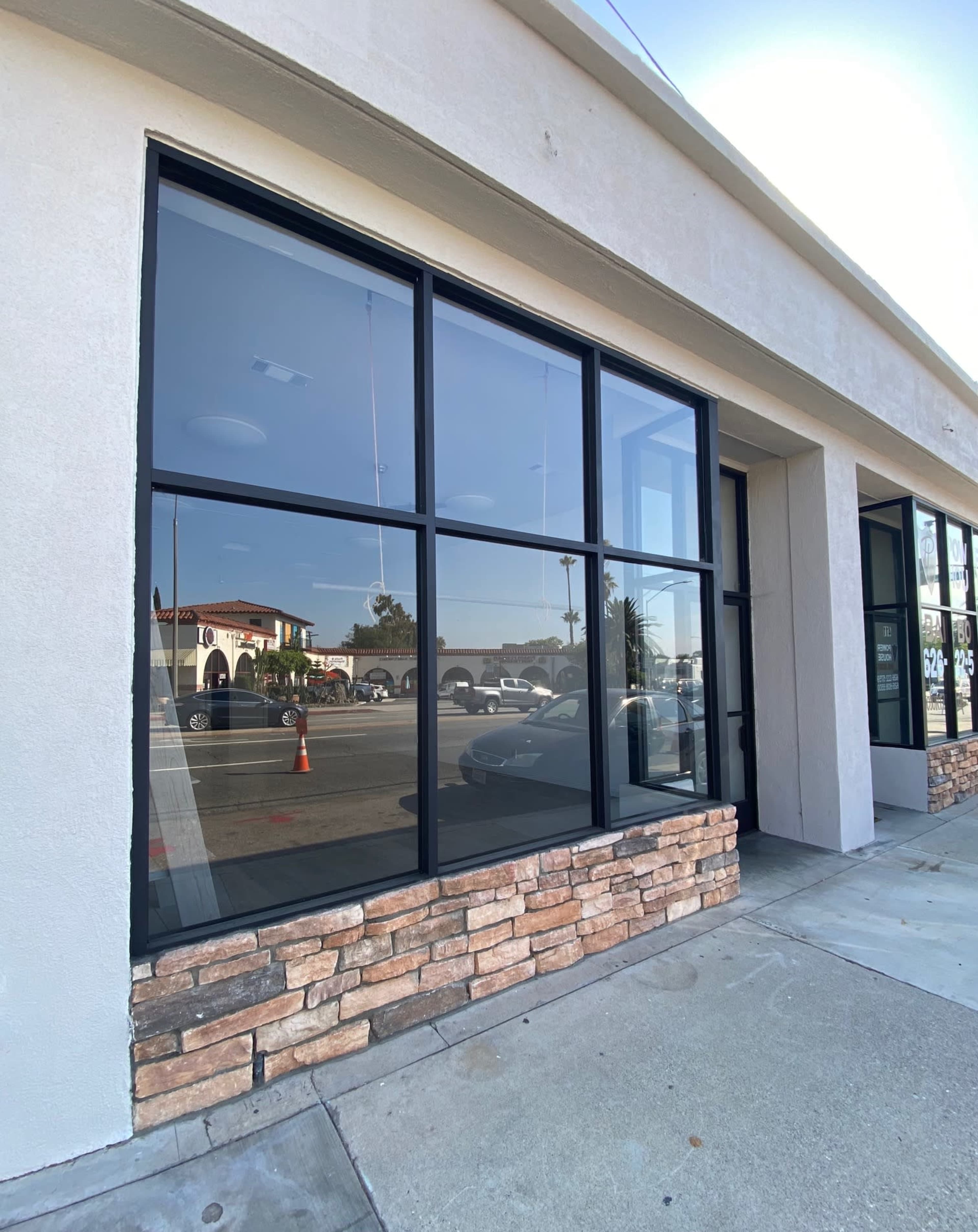 A storefront with large glass windows framed in black and a stone accent at the base, located on a paved sidewalk.