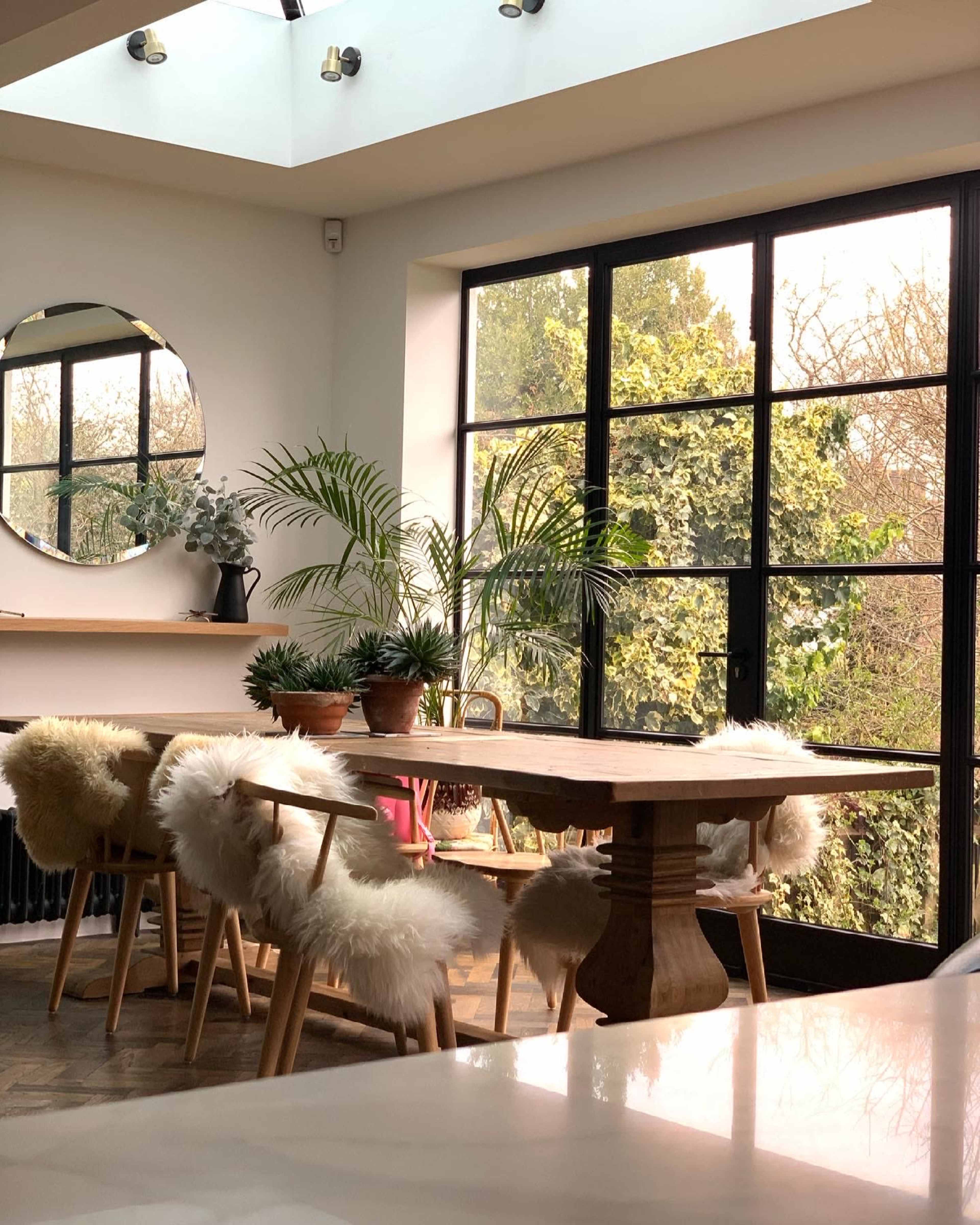 A dining area features a wooden table surrounded by chairs with fur seats, large windows displaying green outdoor foliage, and a circular mirror on the wall.