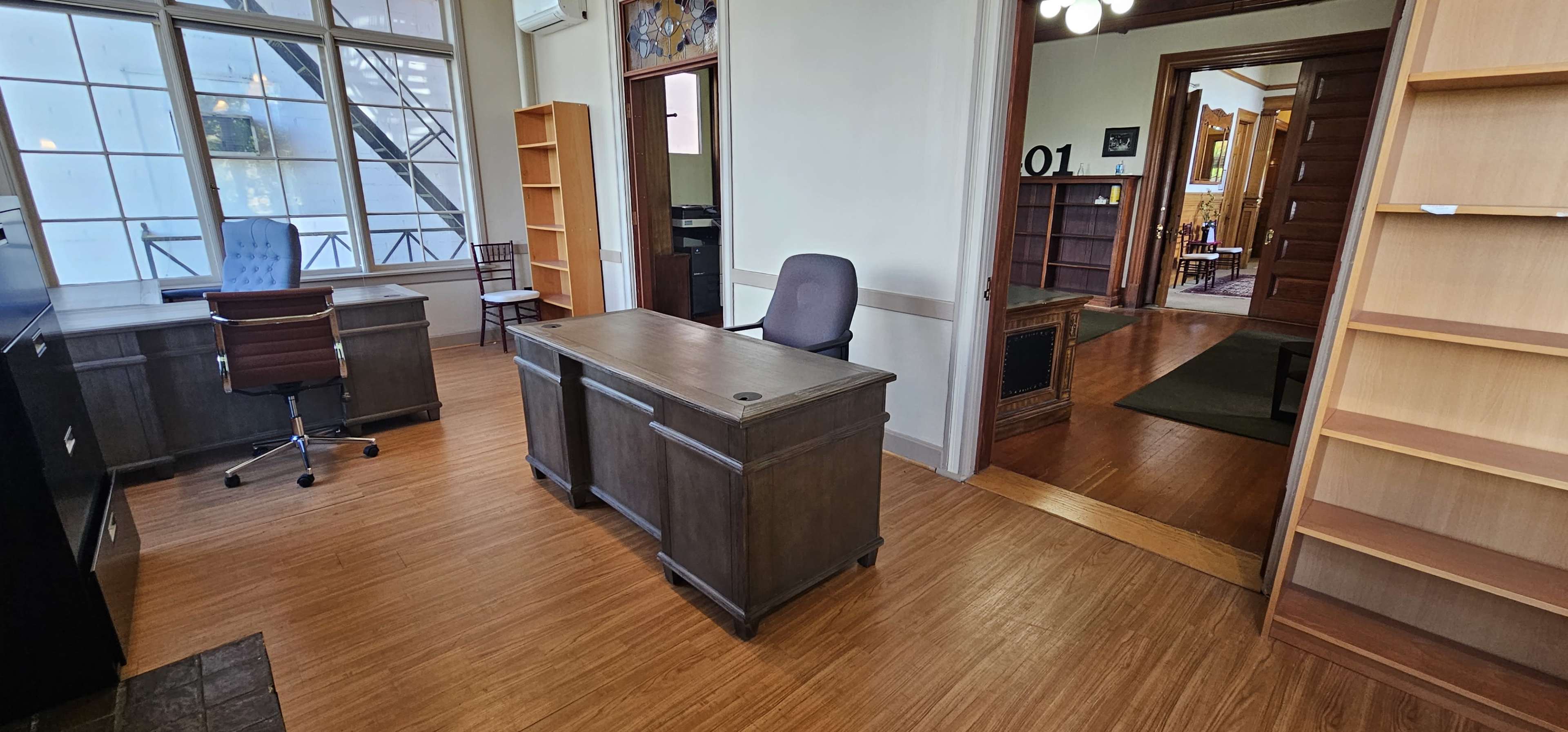 The image depicts a spacious office area featuring a desk, a chair, and shelves, with a doorway leading to another room.