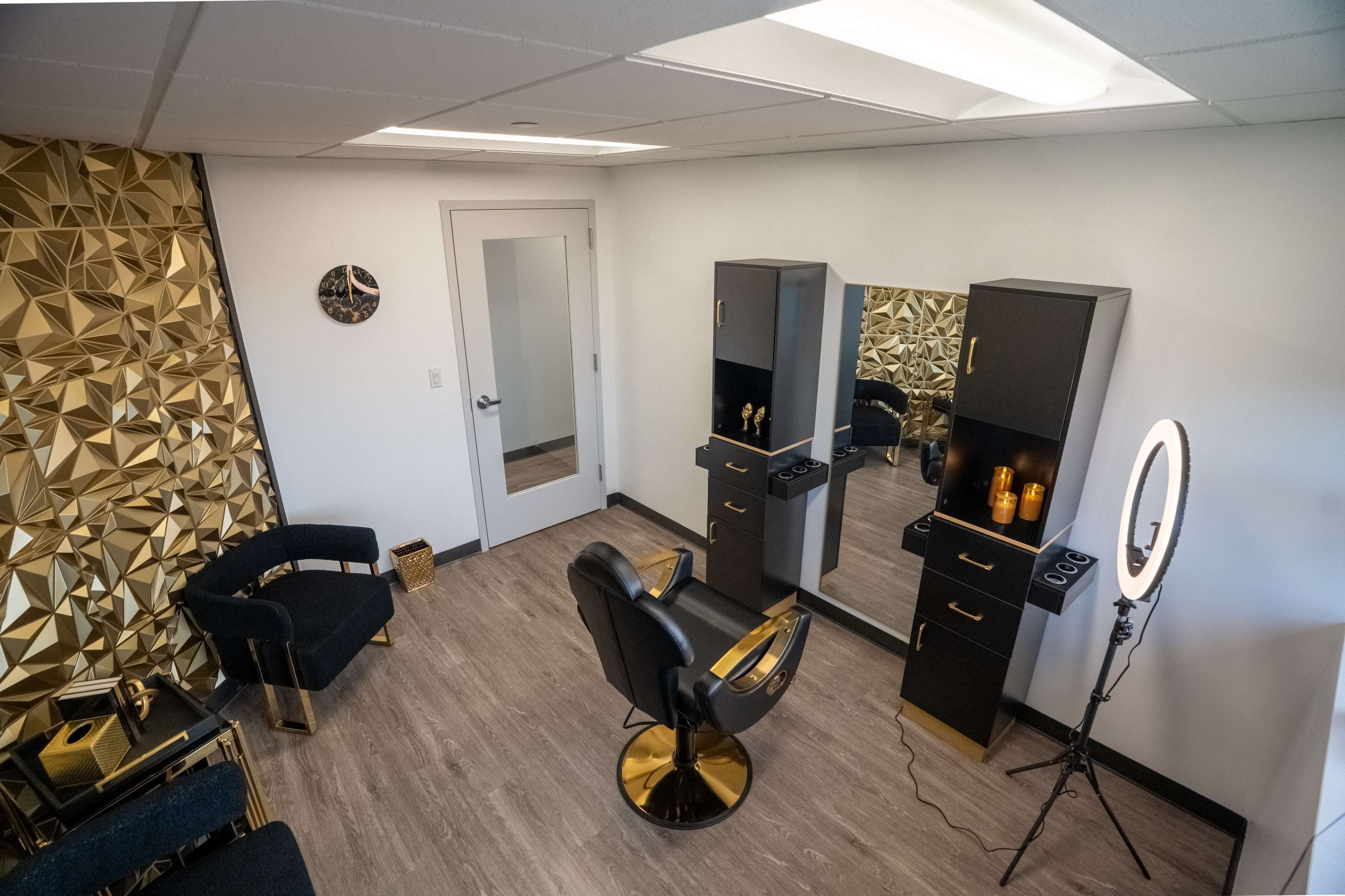 The image shows a modern hair salon interior with a black and gold color theme, featuring a styling chair, mirrors, and a ring light setup.