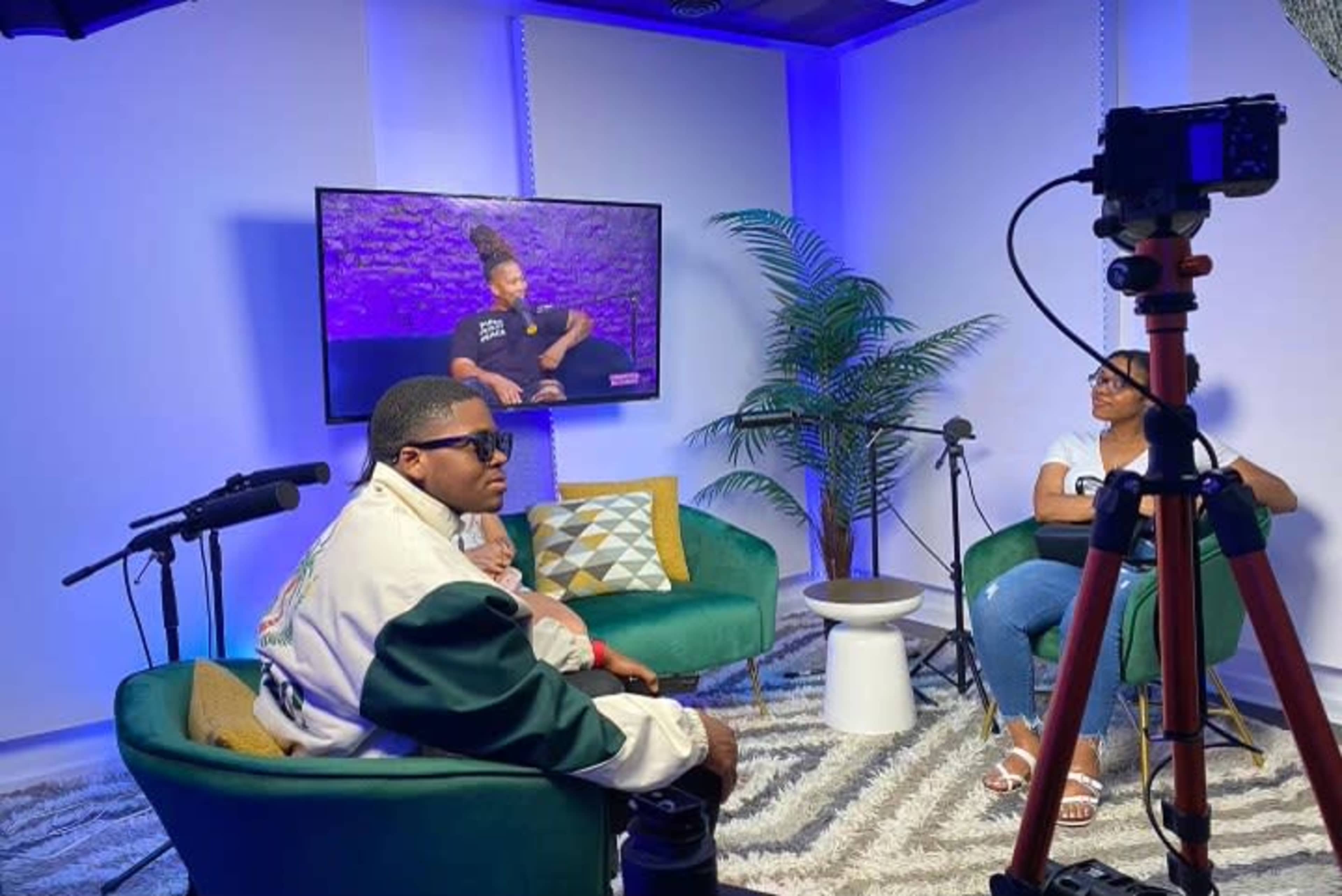 A small studio setup features two people seated on green chairs, with a television displaying a program in the background and a camera on a tripod aimed at them.