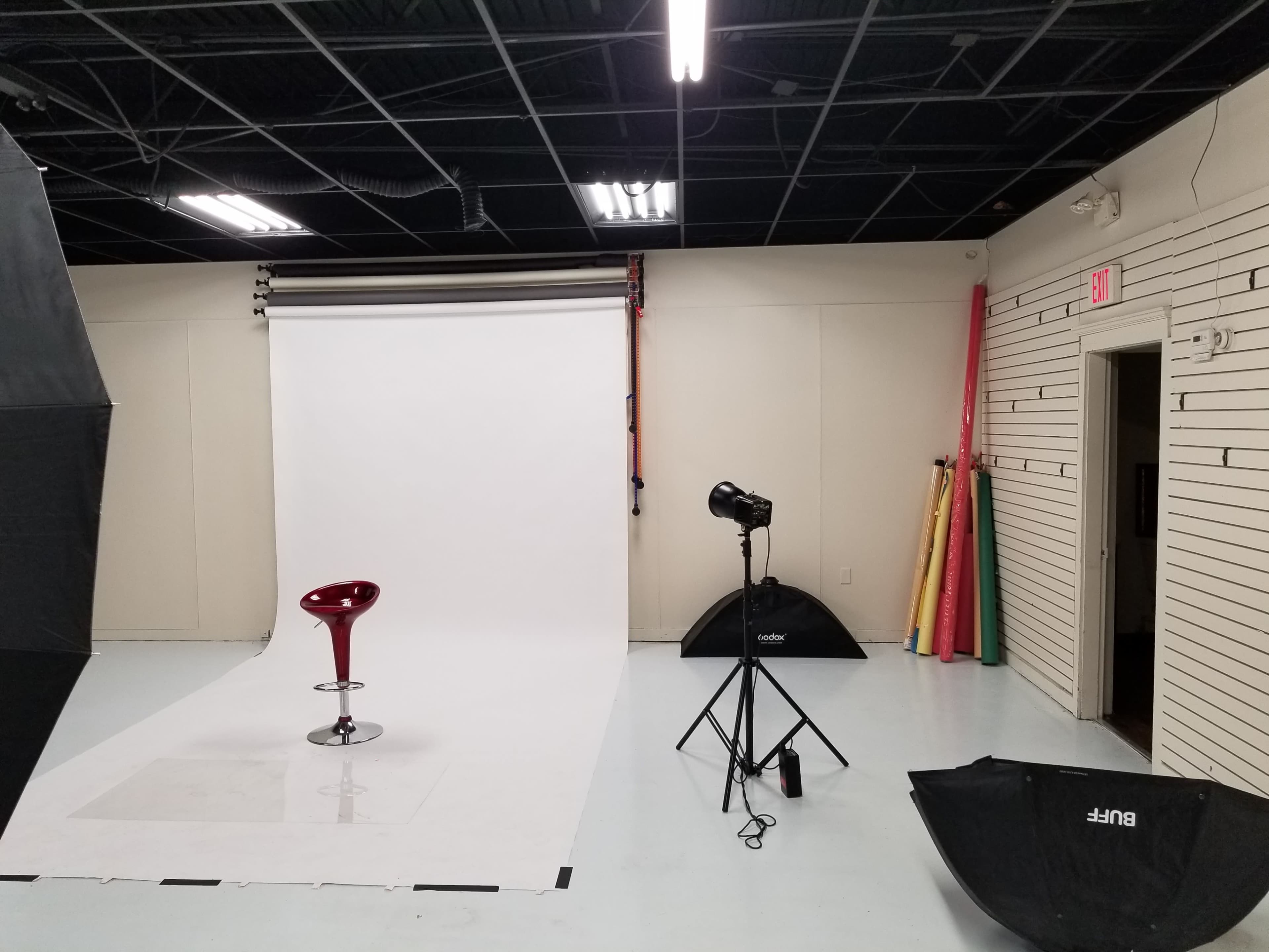 A photography studio with a white backdrop, a red stool, a lighting setup, and several colored backdrops leaning against the wall.
