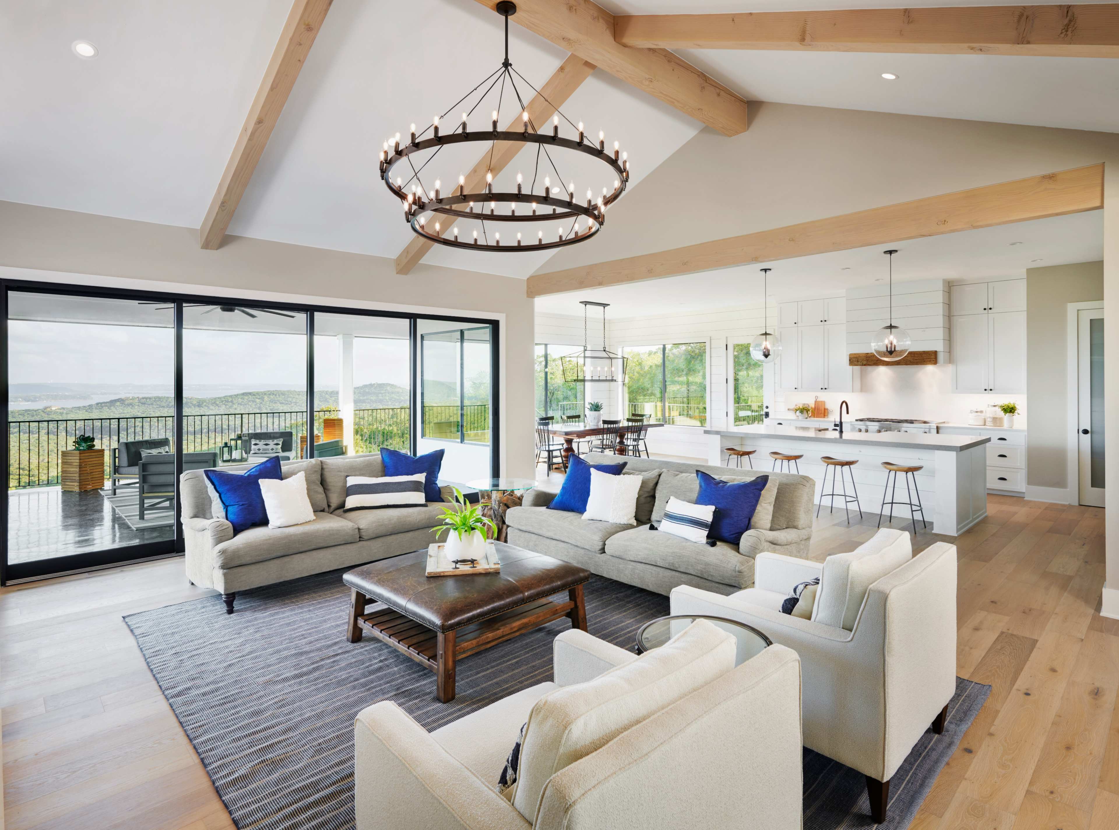 A modern living room with light-colored sofas arranged around a wooden coffee table, large windows providing a view of the outdoors, and a bright kitchen in the background.