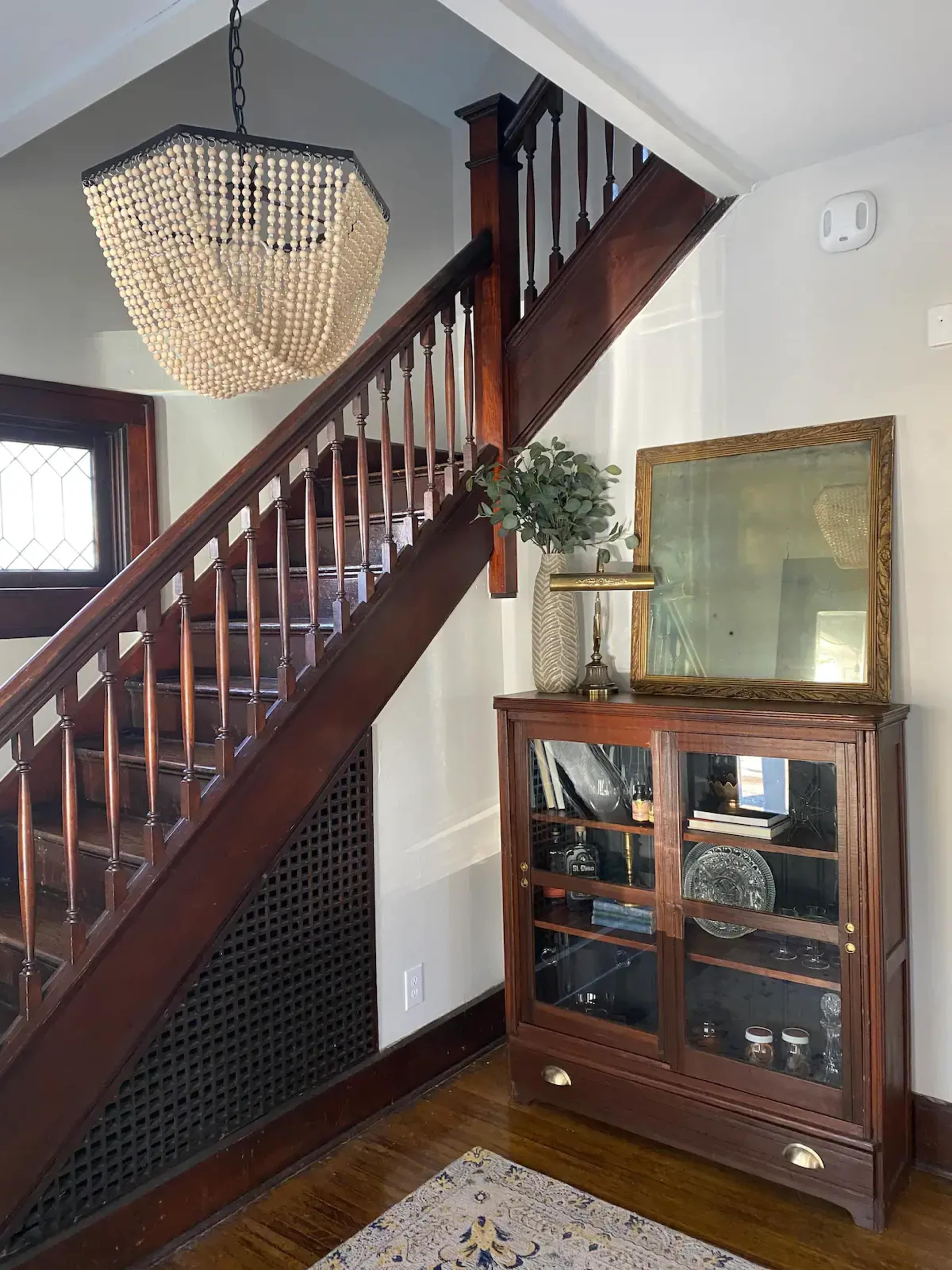 A wooden staircase with a beaded chandelier above, alongside a glass-fronted cabinet displaying various items and a large mirror.