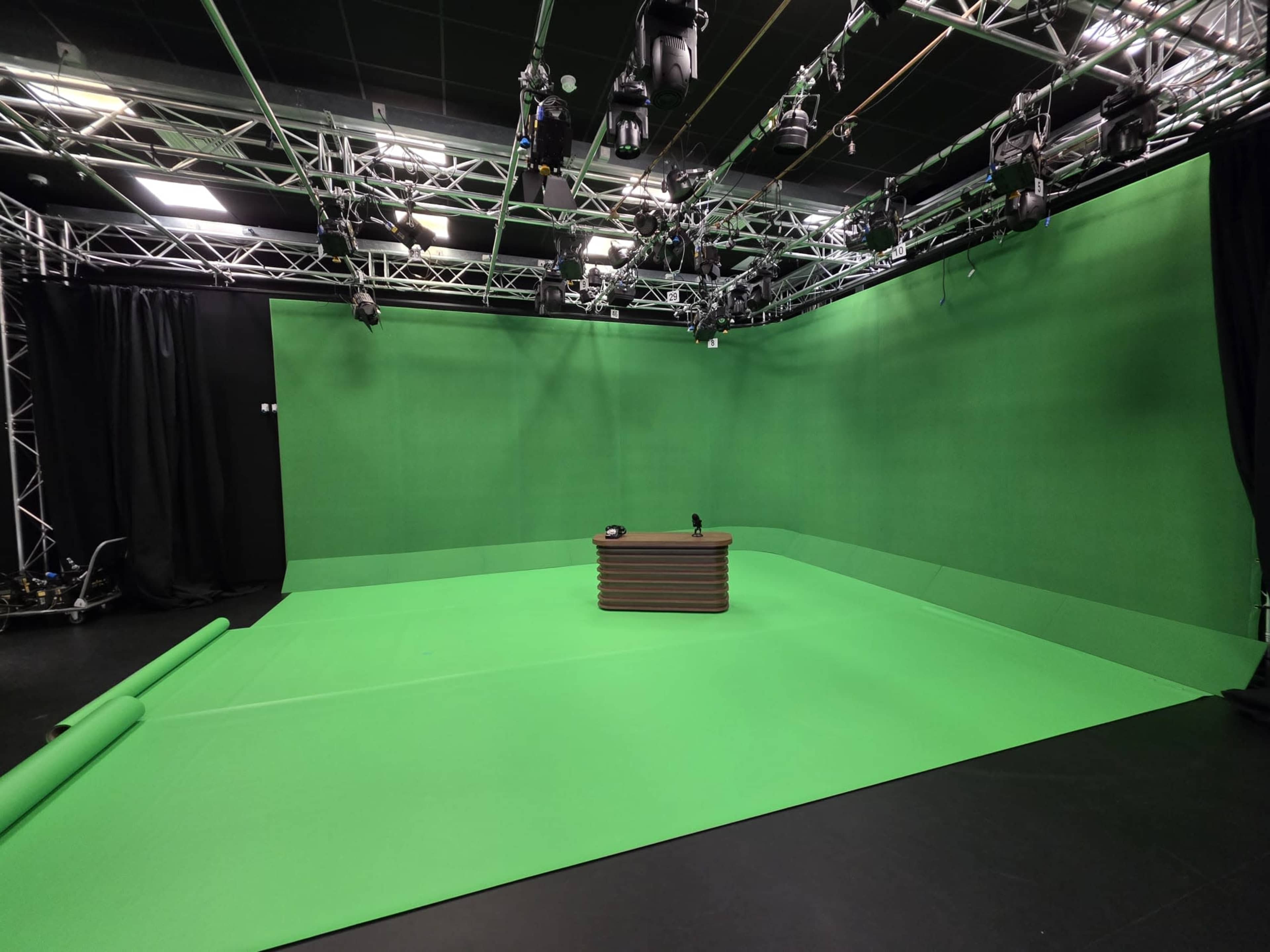 The image shows a green screen studio with a wooden desk in the center, surrounded by black curtains and overhead lighting equipment.
