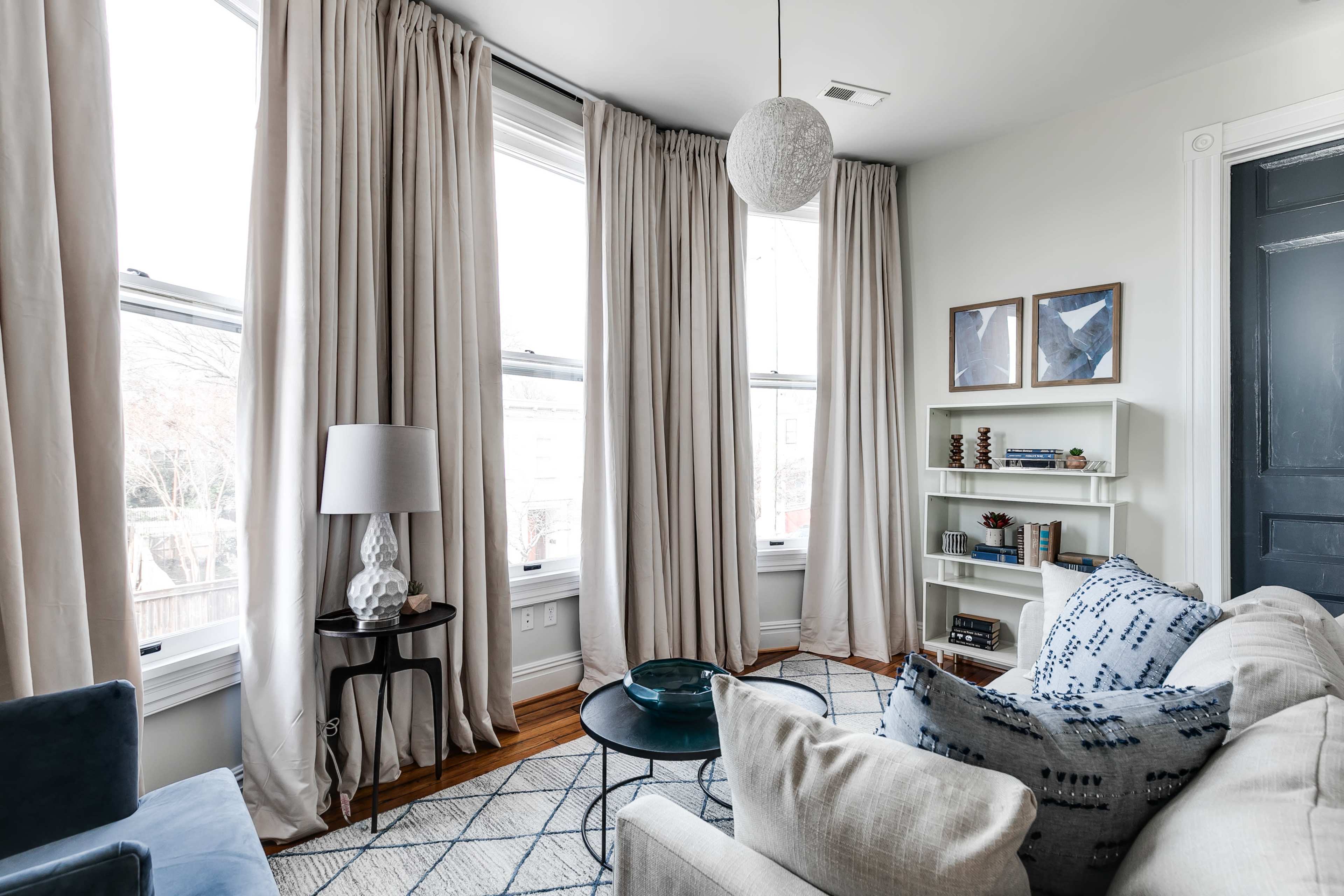 A cozy living room features large windows draped with light-colored curtains, a sectional sofa, a decorative coffee table, and a small shelf filled with books and decor.