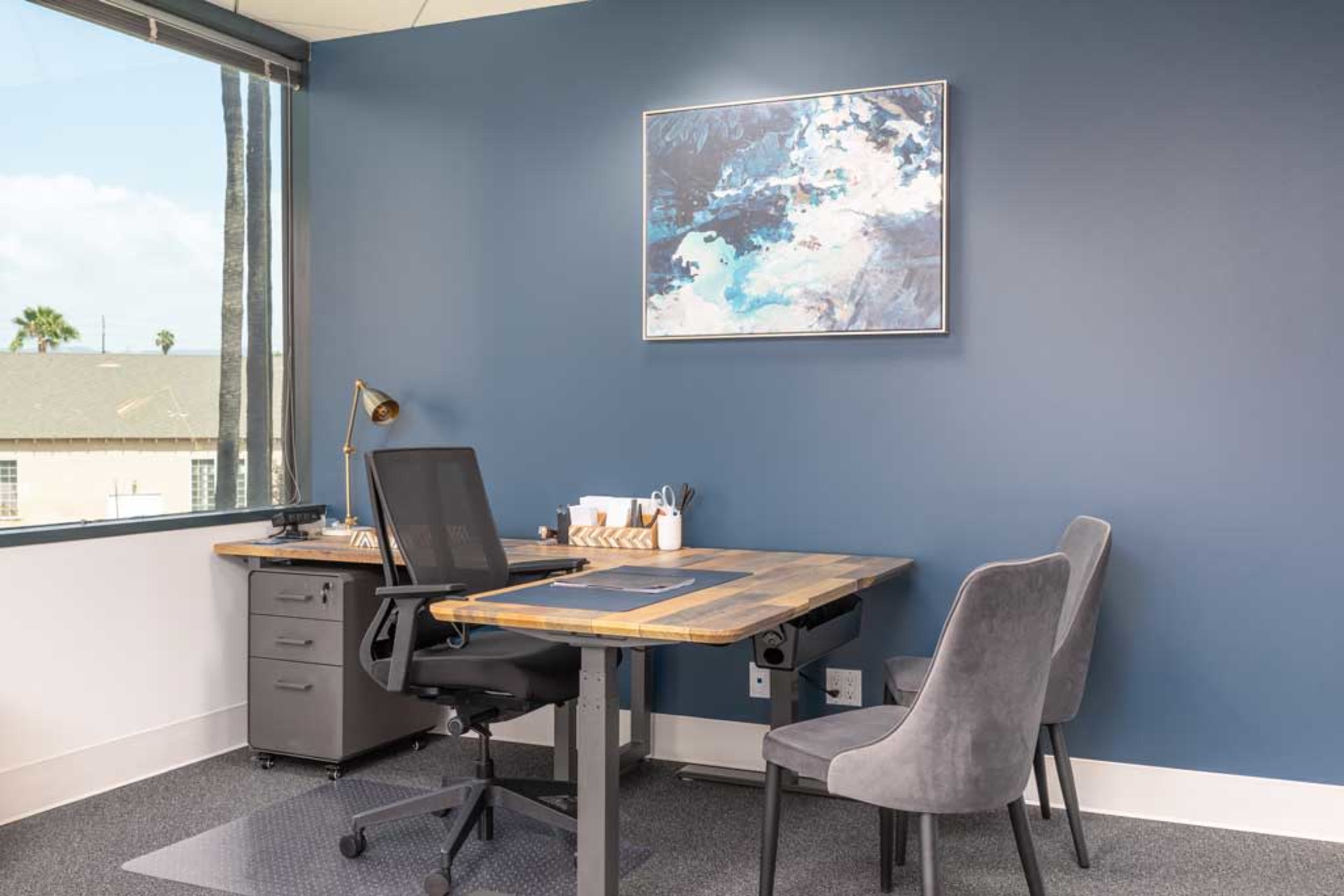A modern office space featuring a wooden desk, a rolling filing cabinet, a gray upholstered chair, and a wall with a blue accent color, adorned with a framed abstract painting.